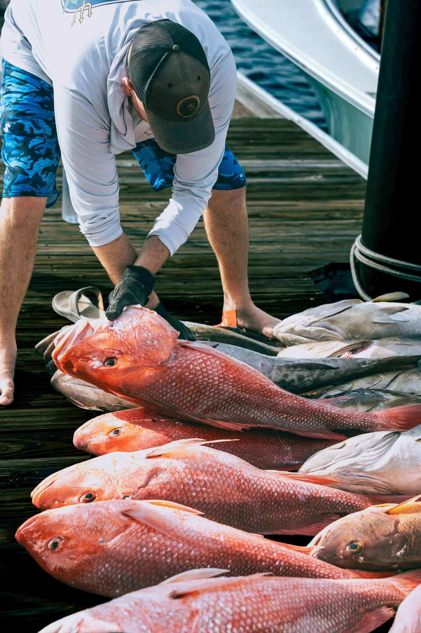 Person in a white long-sleeve shirt and blue camouflage shorts preparing fish on a wooden dock at Point South Marina - Bay Point (Panama City Beach), with a boat nearby.