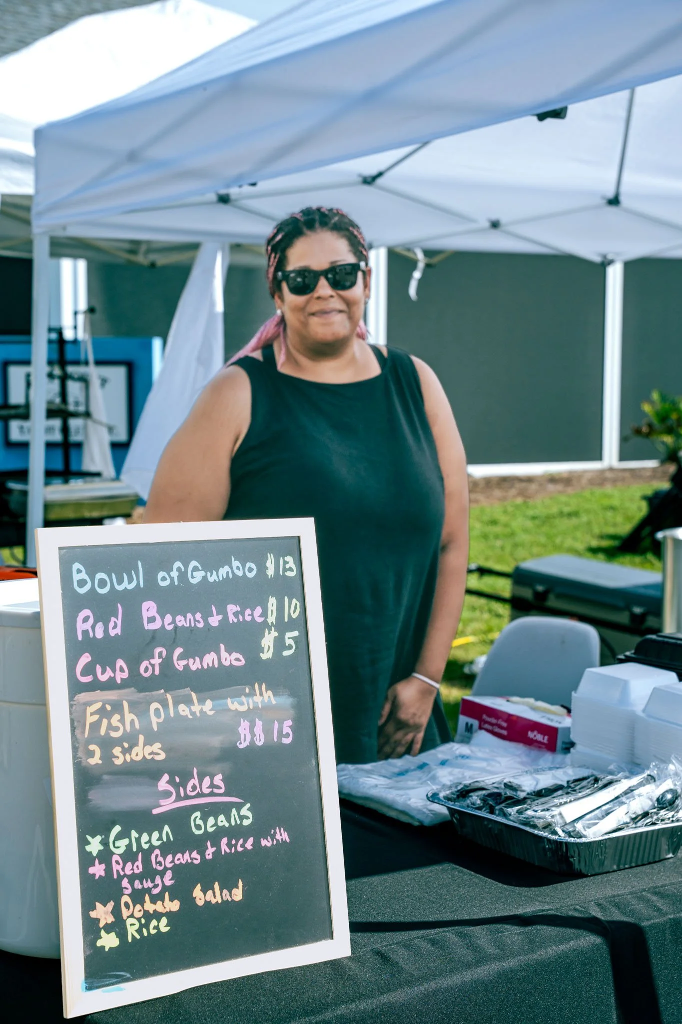 A woman with pink dreadlocks, wearing sunglasses and a black sleeveless shirt, standing behind a table at an outdoor food stall under white tents. There is a chalkboard menu with colorful writing listing food items including gumbo, baked beans, rice,