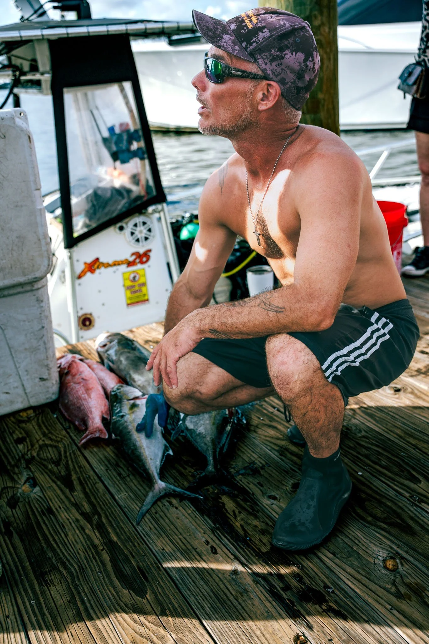 A shirtless man wearing sunglasses, a cap, and shorts, kneeling on a wooden dock next to several fish he caught, with boats in the background.