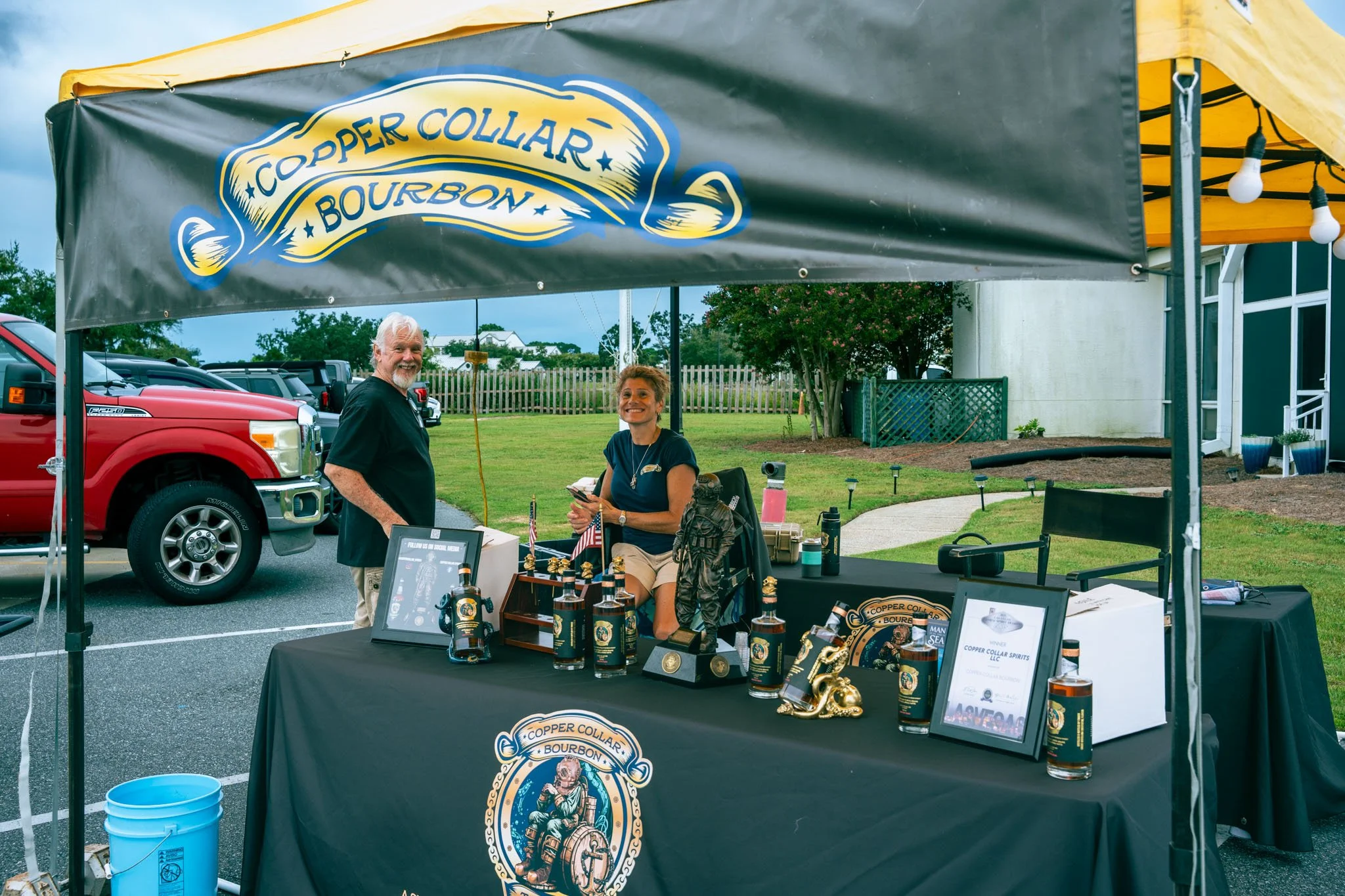 An outdoor booth with a black tablecloth displaying bottles of copper collar bourbon spirit, with two smiling women behind the table, one standing and one sitting, under a canopy with string lights. A large banner above the booth reads 'Copper Collar