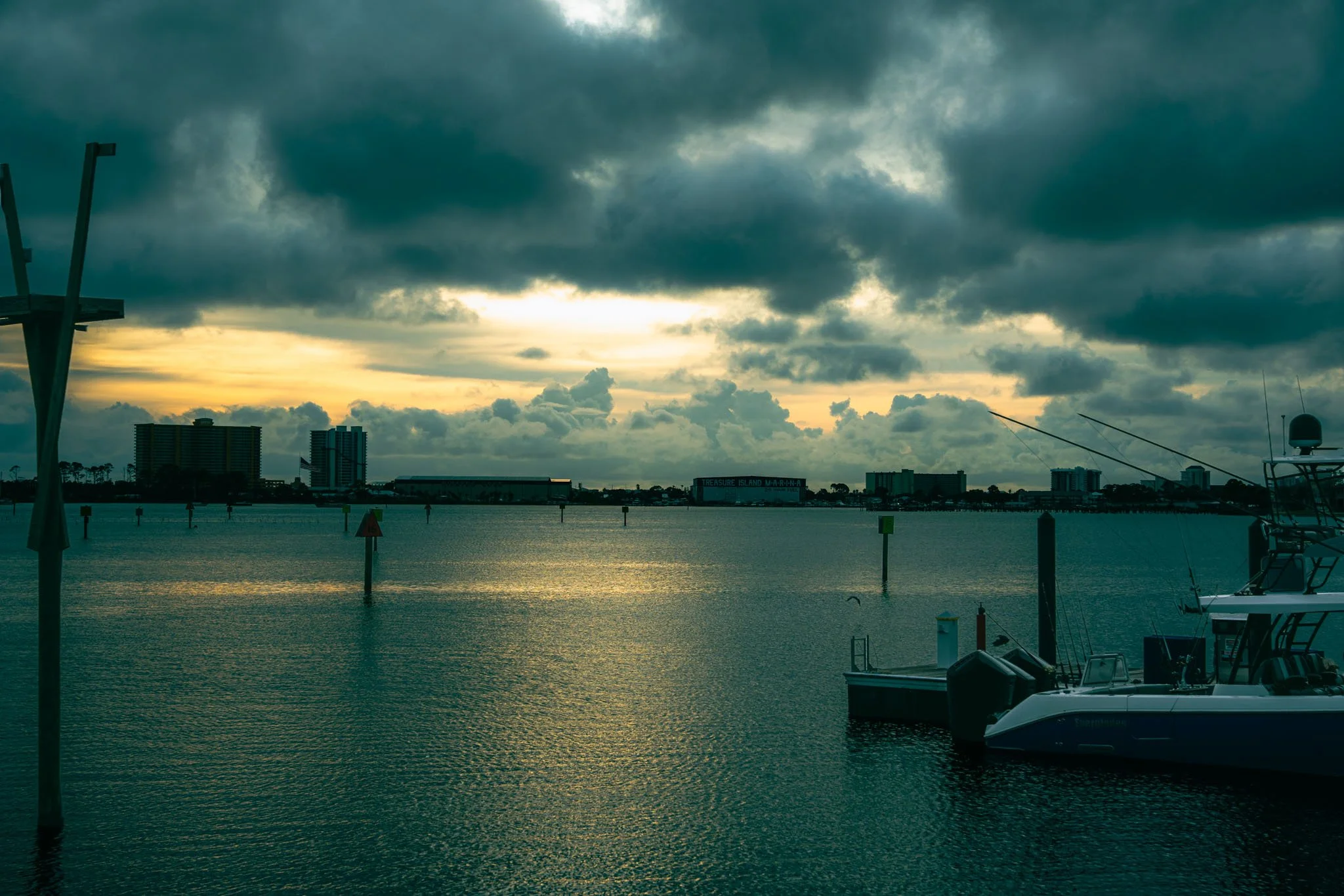 Sunset over a harbor with boats docked, city buildings in the background, and dark clouds in the sky.