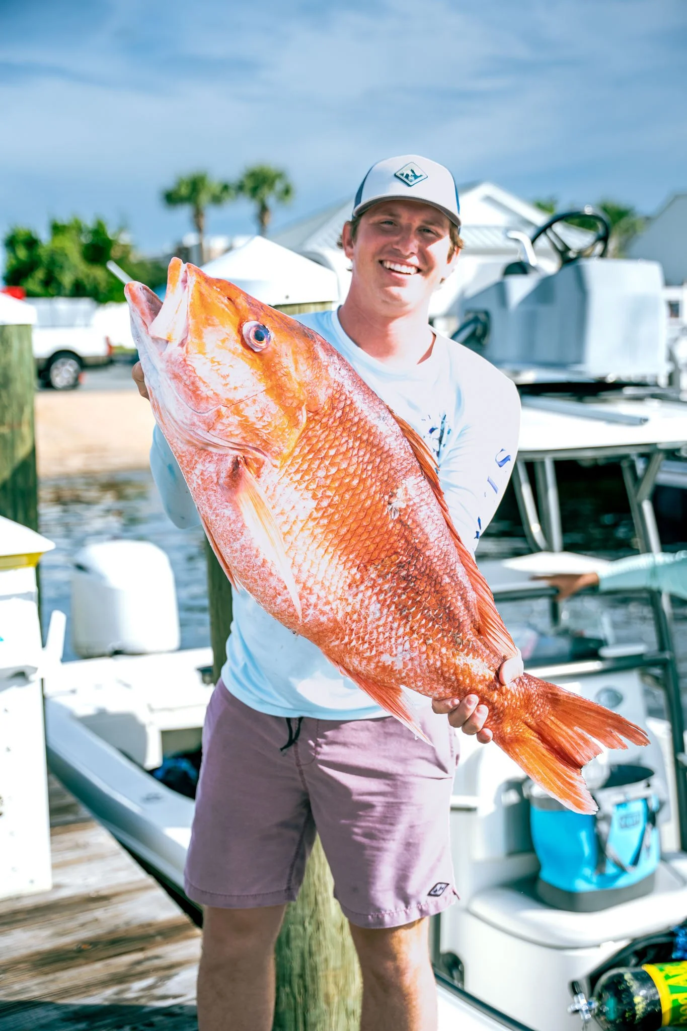 A smiling man wearing a baseball cap and shorts, holding a large red fish on a dock at Point South Marina - Bay Point (Panama City Beach) with boats, palm trees, and a blue sky in the background.