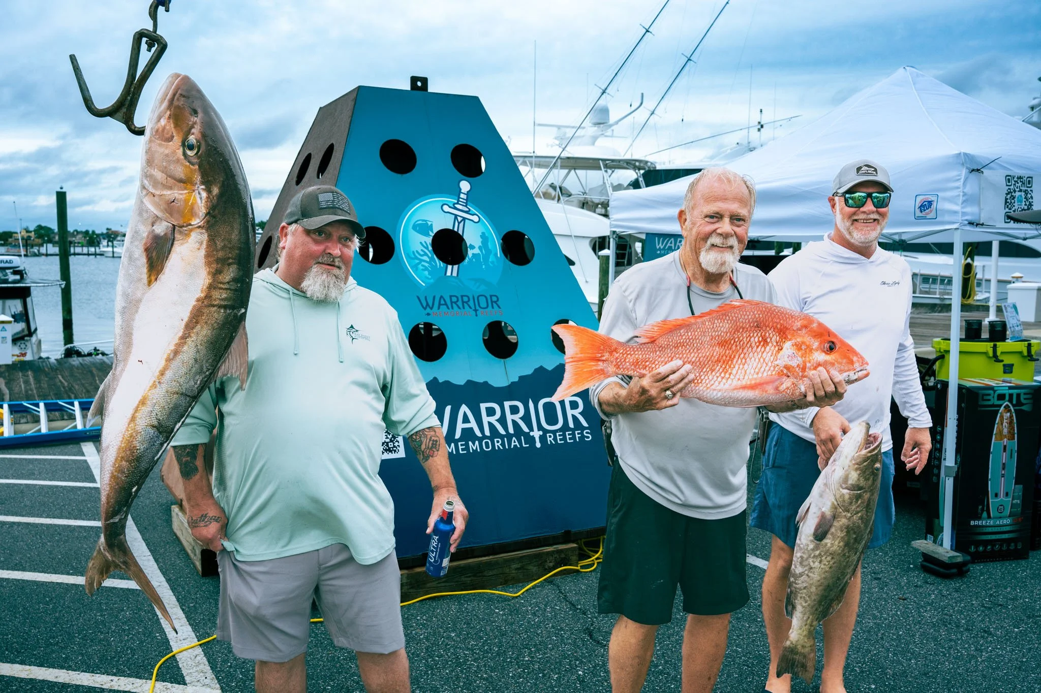 Three men holding large fish at Point South Marina - Bay Point (Panama City Beach), with boats and a blue sign that reads 'Warrior Memorial Reefs' in the background.