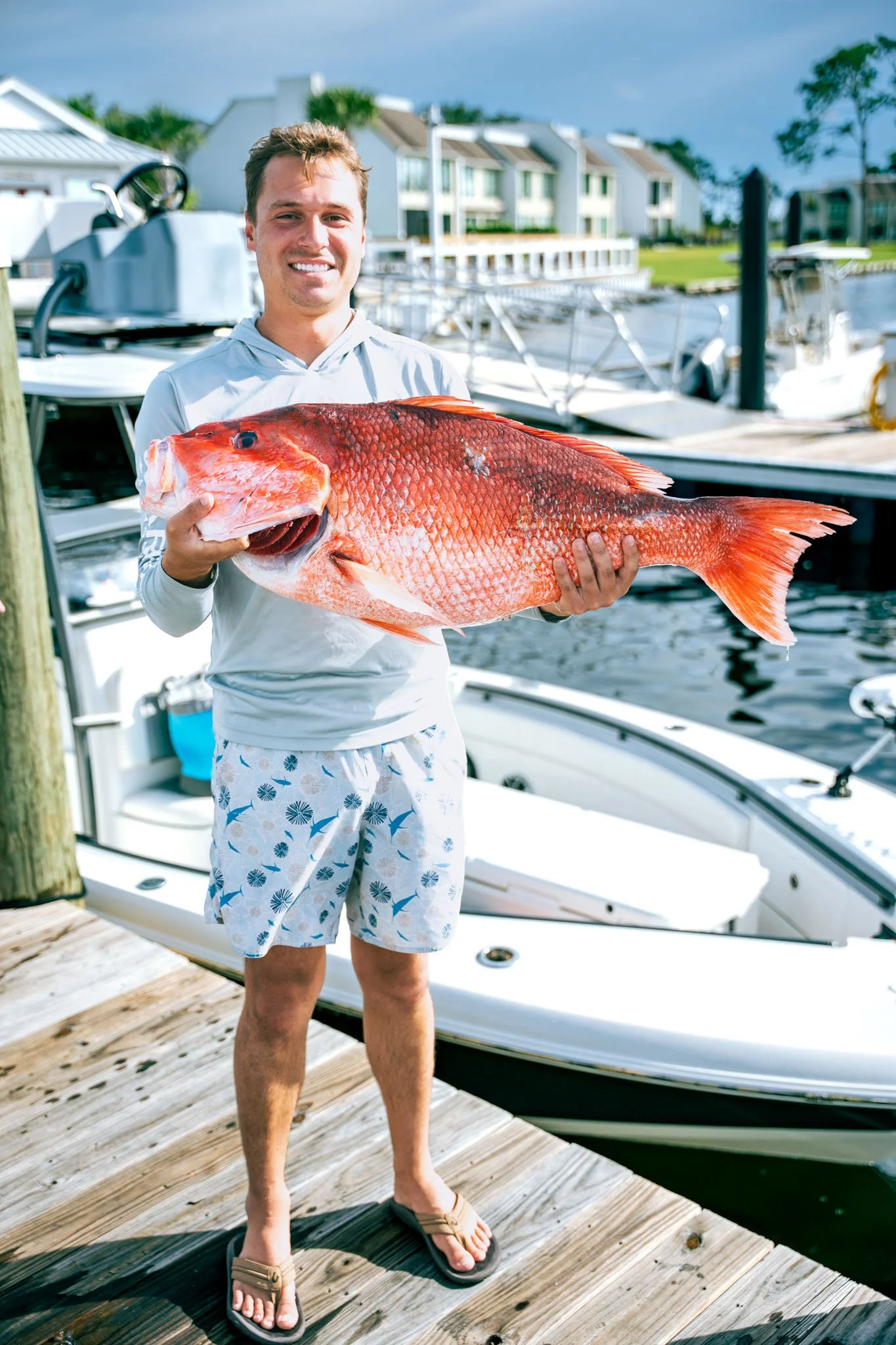 A man standing on a wooden dock at Point South Marina - Bay Point (Panama City Beach), holding a large red fish, near boats by a body of water with houses and trees in the background on a sunny day.
