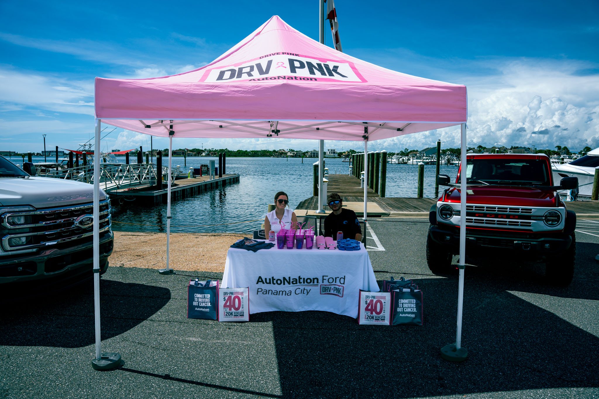 A pink Canopy with the 'DRV PNK' logo and 'AutoNation' branding, set up on a parking lot near a waterfront with boats and docks. Two people sit behind a table with pink water bottles and promotional materials celebrating AutoNation's Cancer Awareness