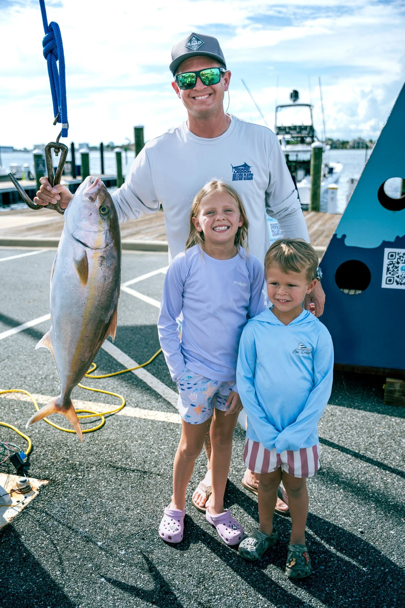 A man holding a large fish on a dock with two children standing next to him, smiling. The man is wearing sunglasses and a cap, and the children are in casual clothing. There are boats and water in the background.
