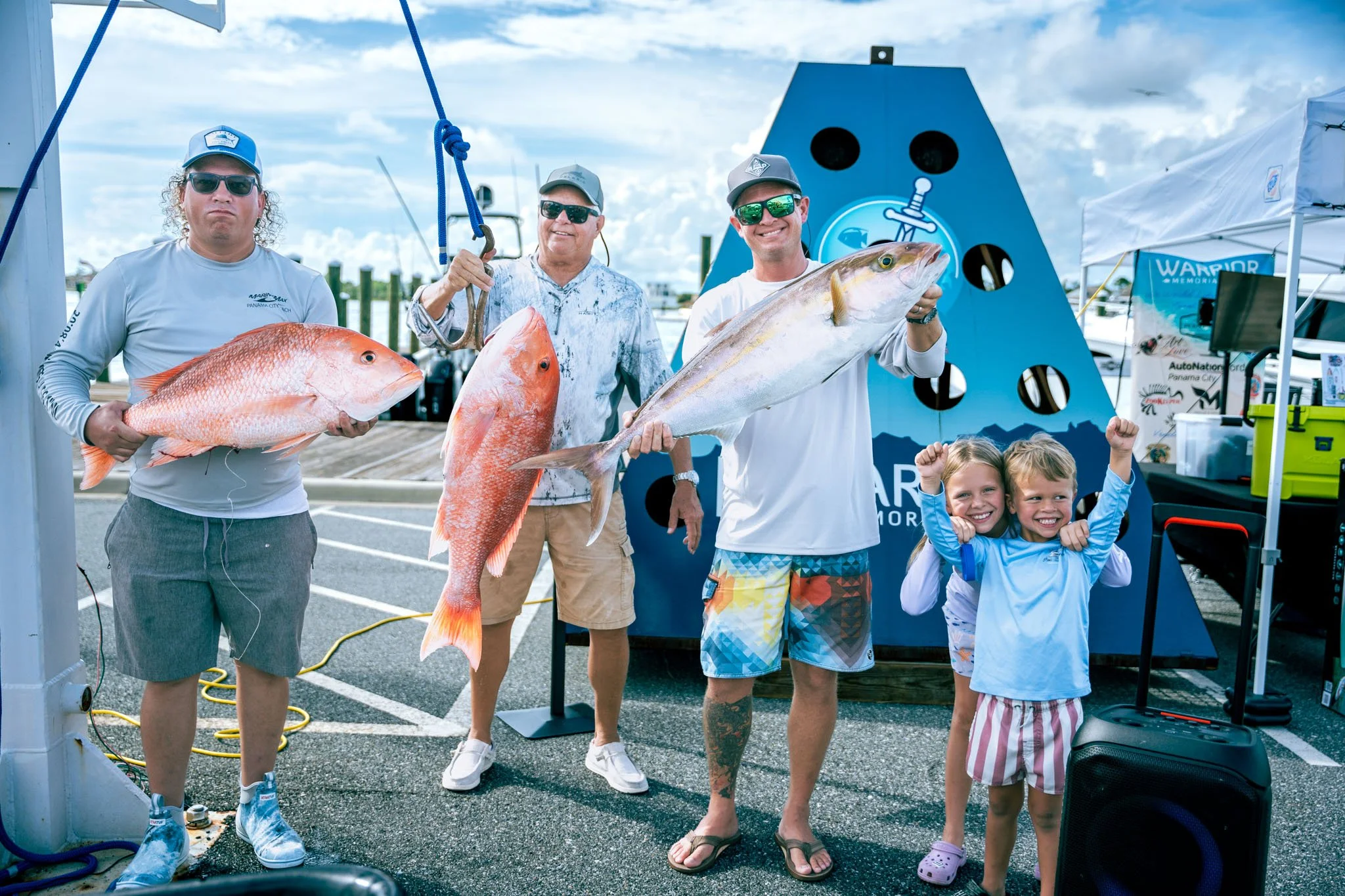 Three men and two young girls pose with large fish they caught, standing in front of fishing gear and a large blue shark silhouette at Point South Marina - Bay Point (Panama City Beach).