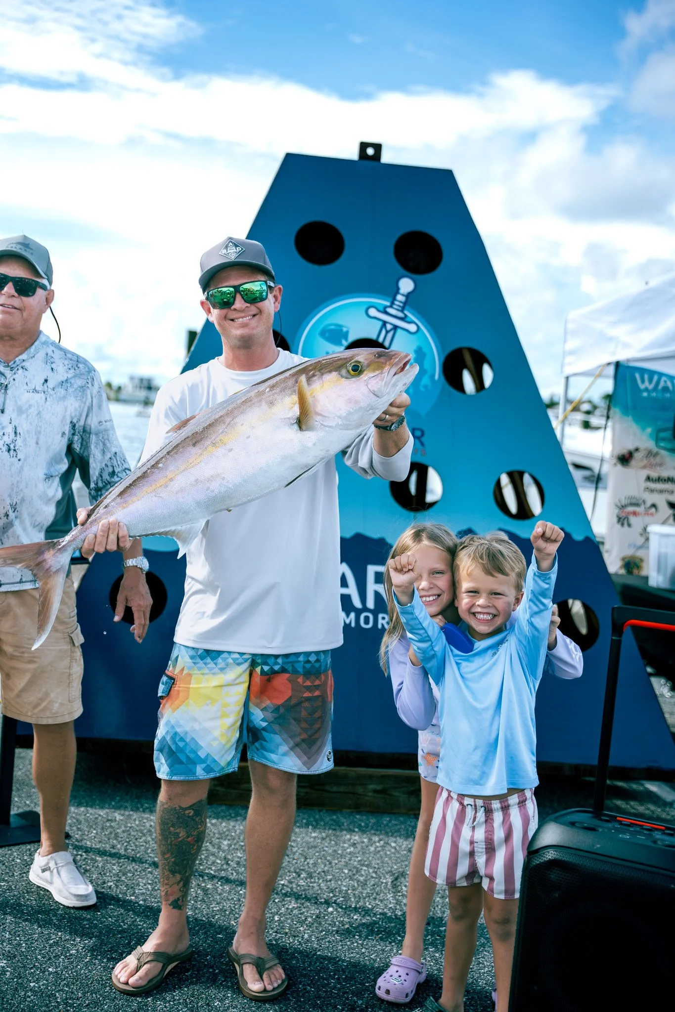 Two children smiling and flexing their muscles while standing in front of a man holding a large fish, with a blue background and logo, at an outdoor event at Point South Marina - Bay Point (Panama City Beach).