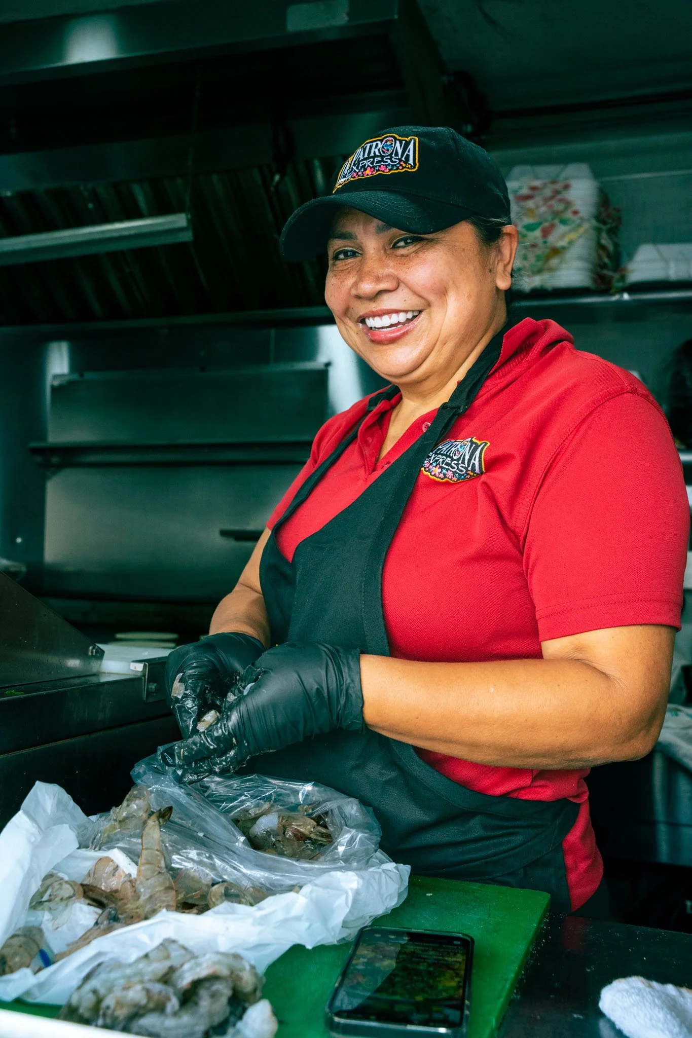 Smiling woman in black cap and red shirt preparing seafood at a food stall, with seafood and a cellphone on the green cutting board.