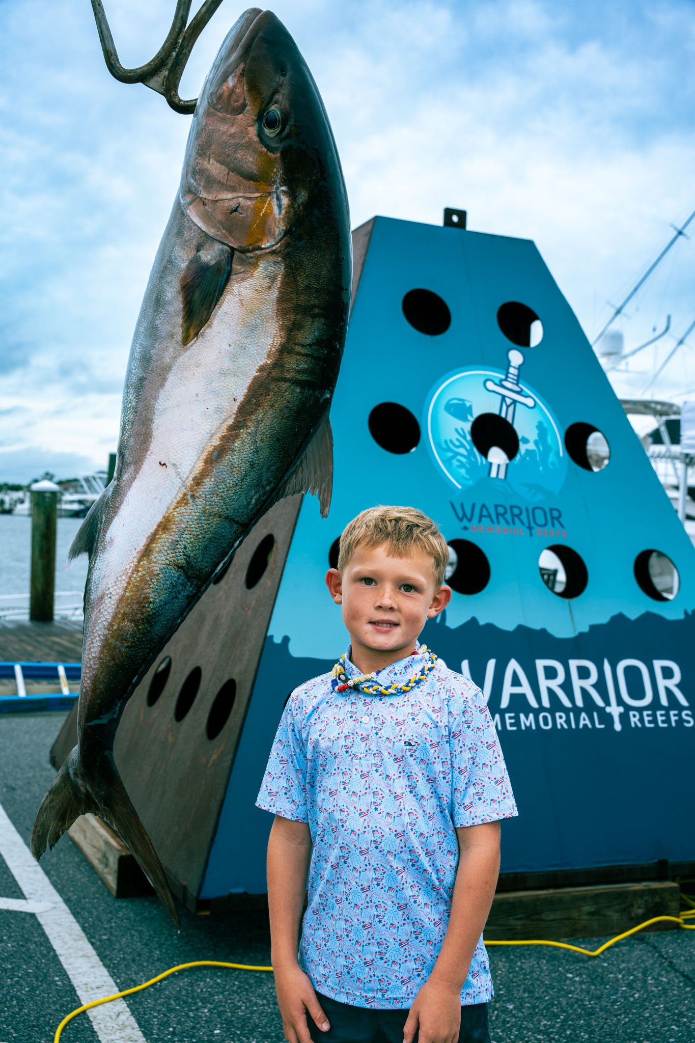 A young boy standing in front of a large fish sculpture at Warrior Memorial Reef. The sculpture features a fish and a blue rectangular sign with black holes, and the boy is wearing a light blue shirt with a pattern and a colorful necklace.