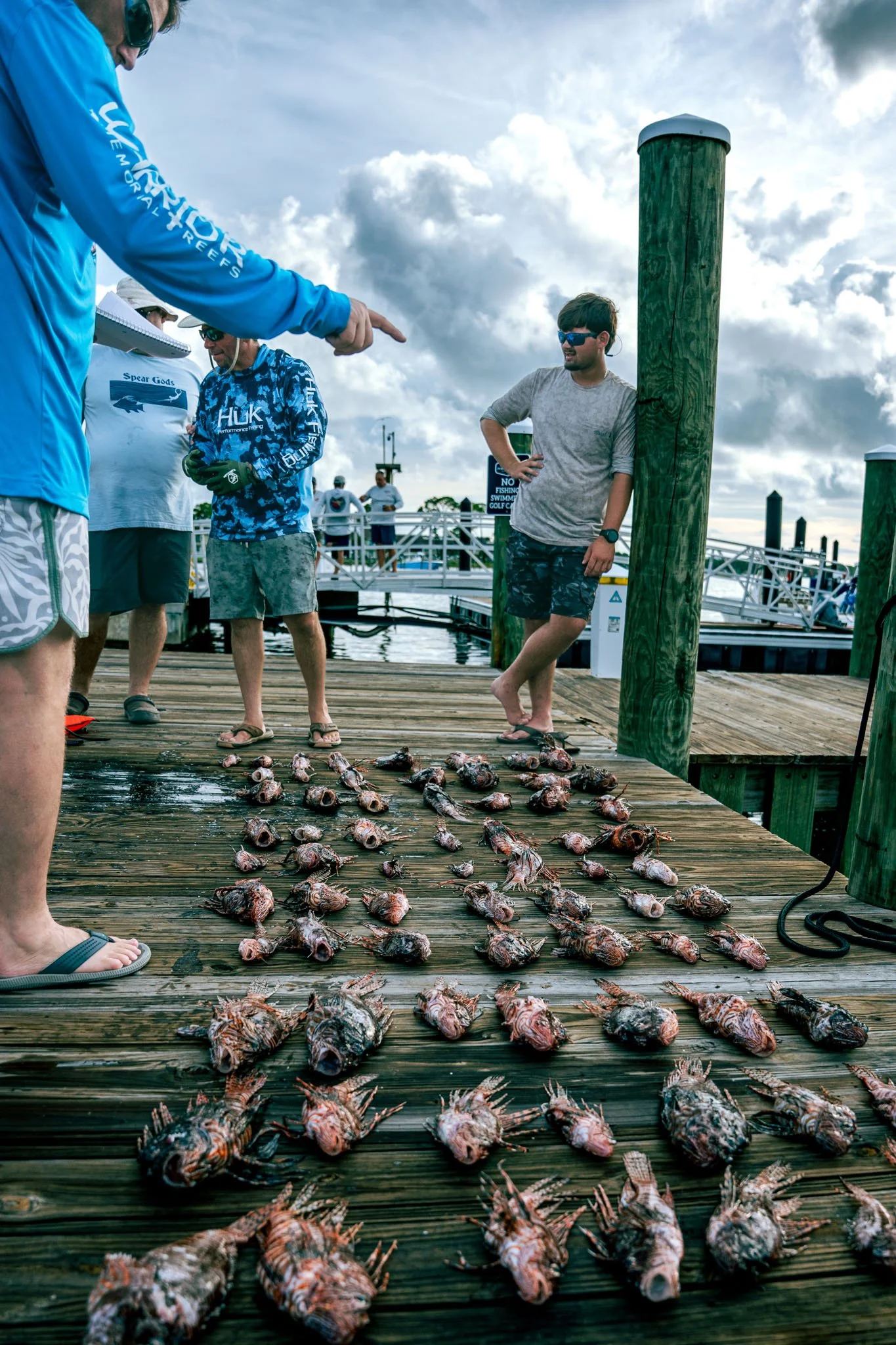 Group of people on a wooden dock examining and counting fish laid out on the dock, with others in the background near boats and a sign.