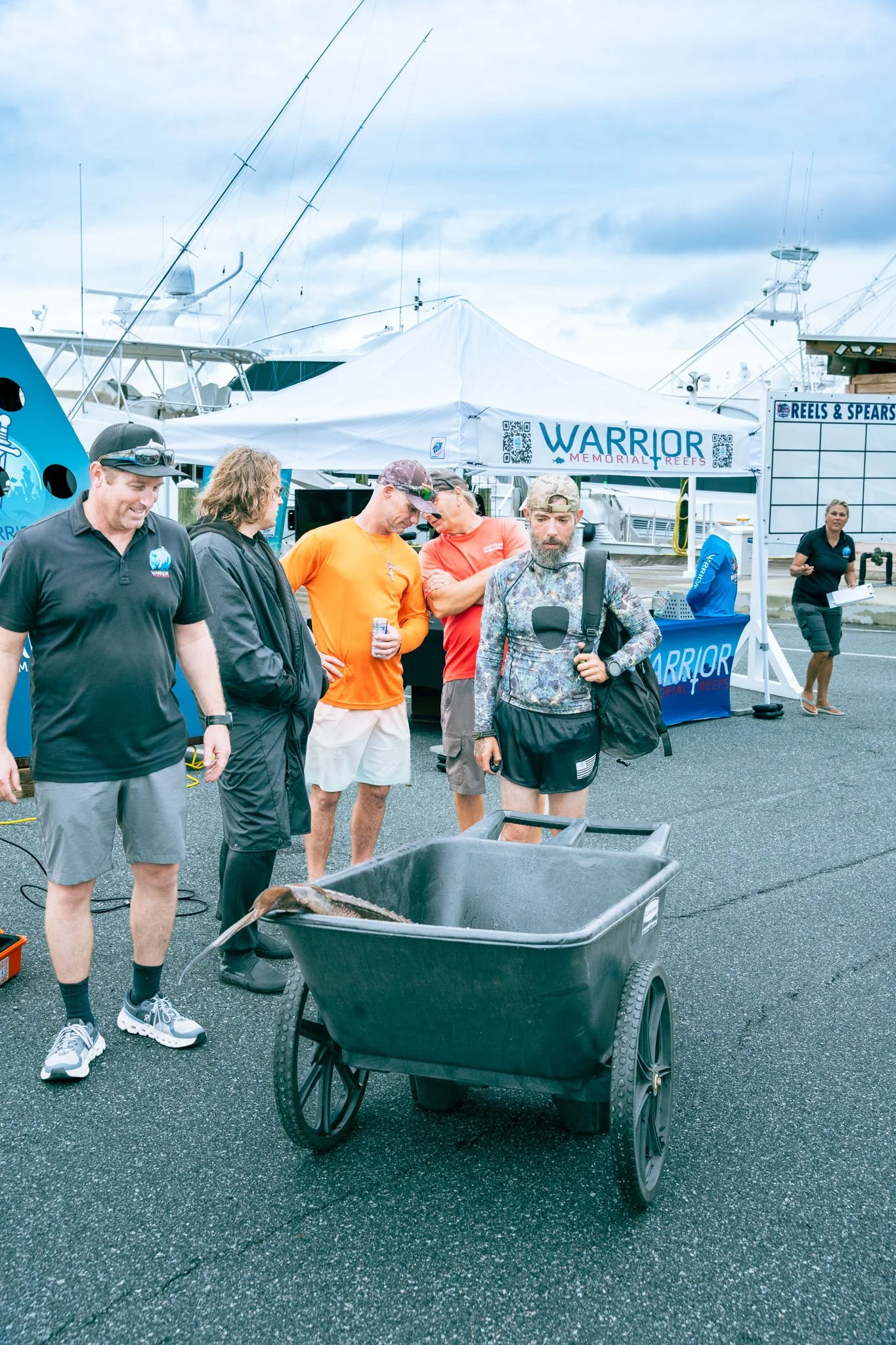 Group of people gathered at a marina event, with boats and yachts in the background, and a white tent labeled "Warrior Memorial Reefs." A man with a backpack and beard stands in front of a black trash bin with a large fish inside. Others are engaged 
