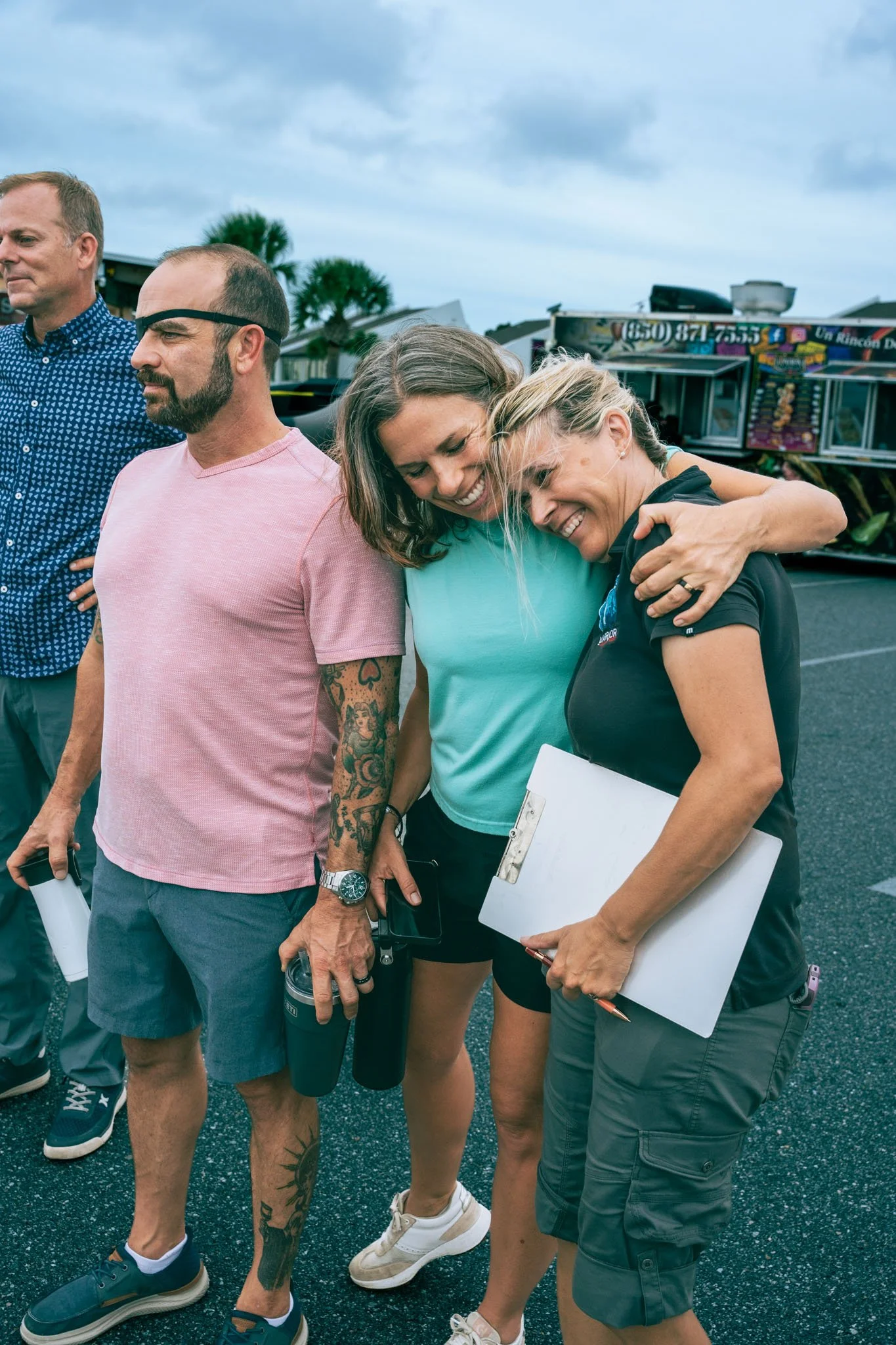 Four people standing close together outdoors at Point South Marina - Bay Point on a cloudy day, with a food truck and palm trees in the background. Two women are smiling and embracing, two men stand nearby, one holding a clipboard and a thermos.
