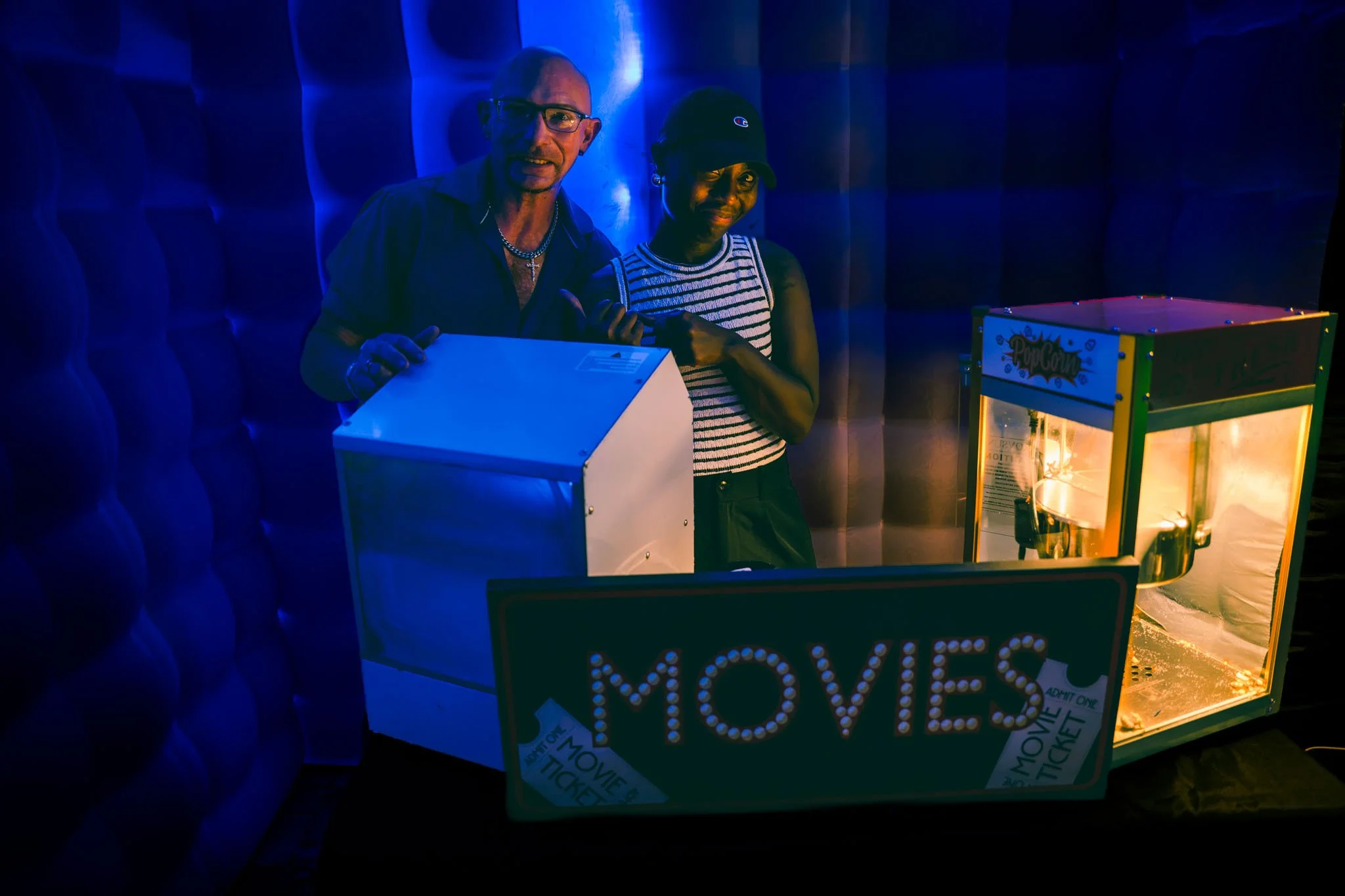 Two people standing beside a popcorn machine and a ticket sign at a venue with dark, blue lighting.