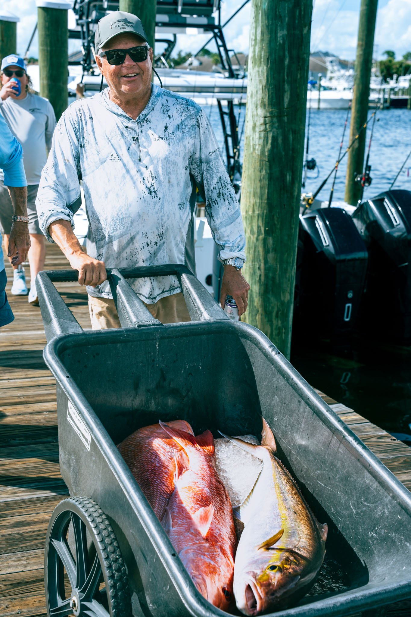 A man wearing sunglasses, a light-colored shirt, a cap, and a watch is smiling while holding a handle on a cart filled with a large fish, with a dock and several boats in the background.