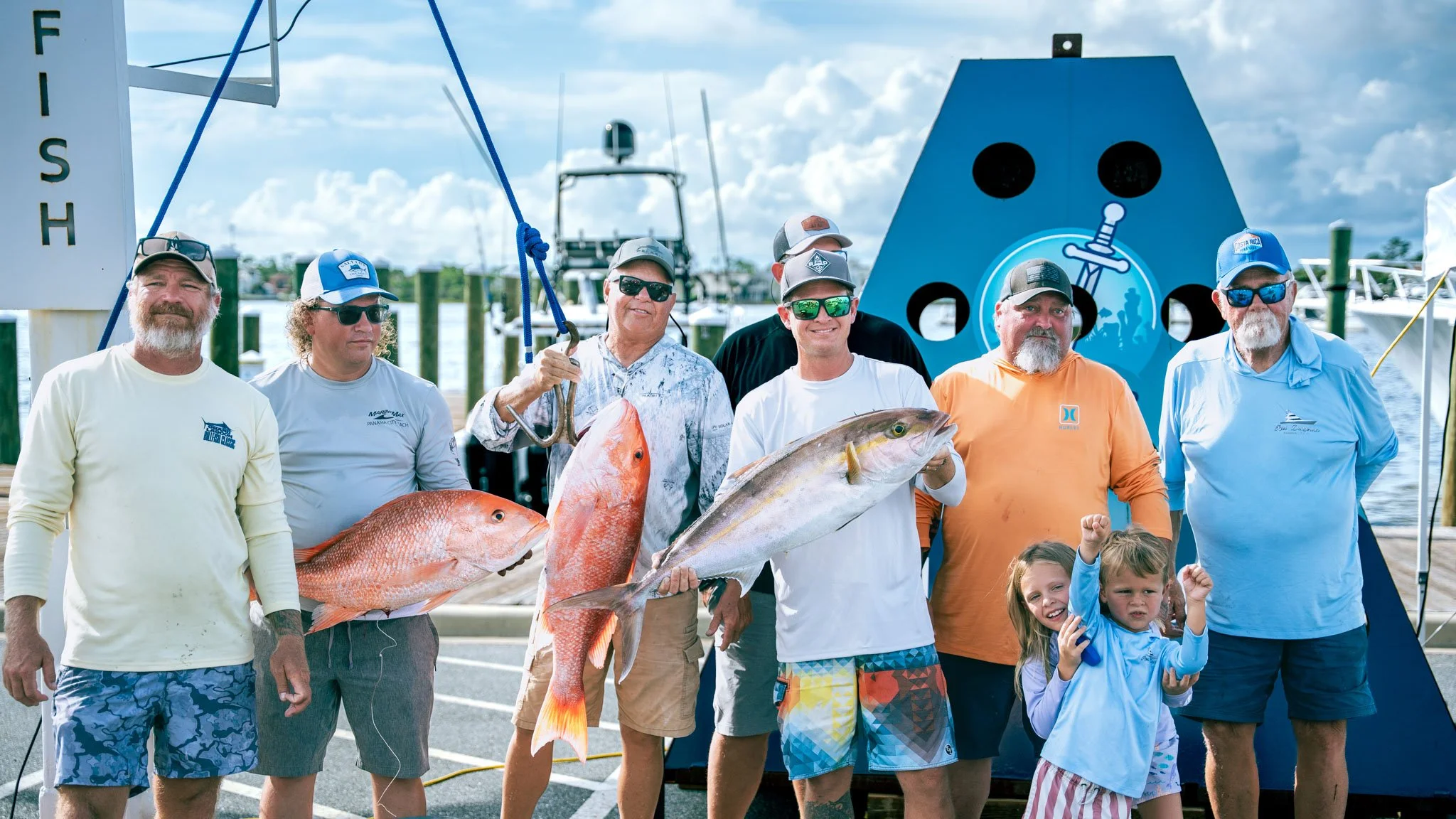 A group of people, including children, standing in front of a boat dock with several boats in the background. They are proudly displaying large fish they caught, with one man holding a red snapper and another holding a yellowtail. The group is smilin