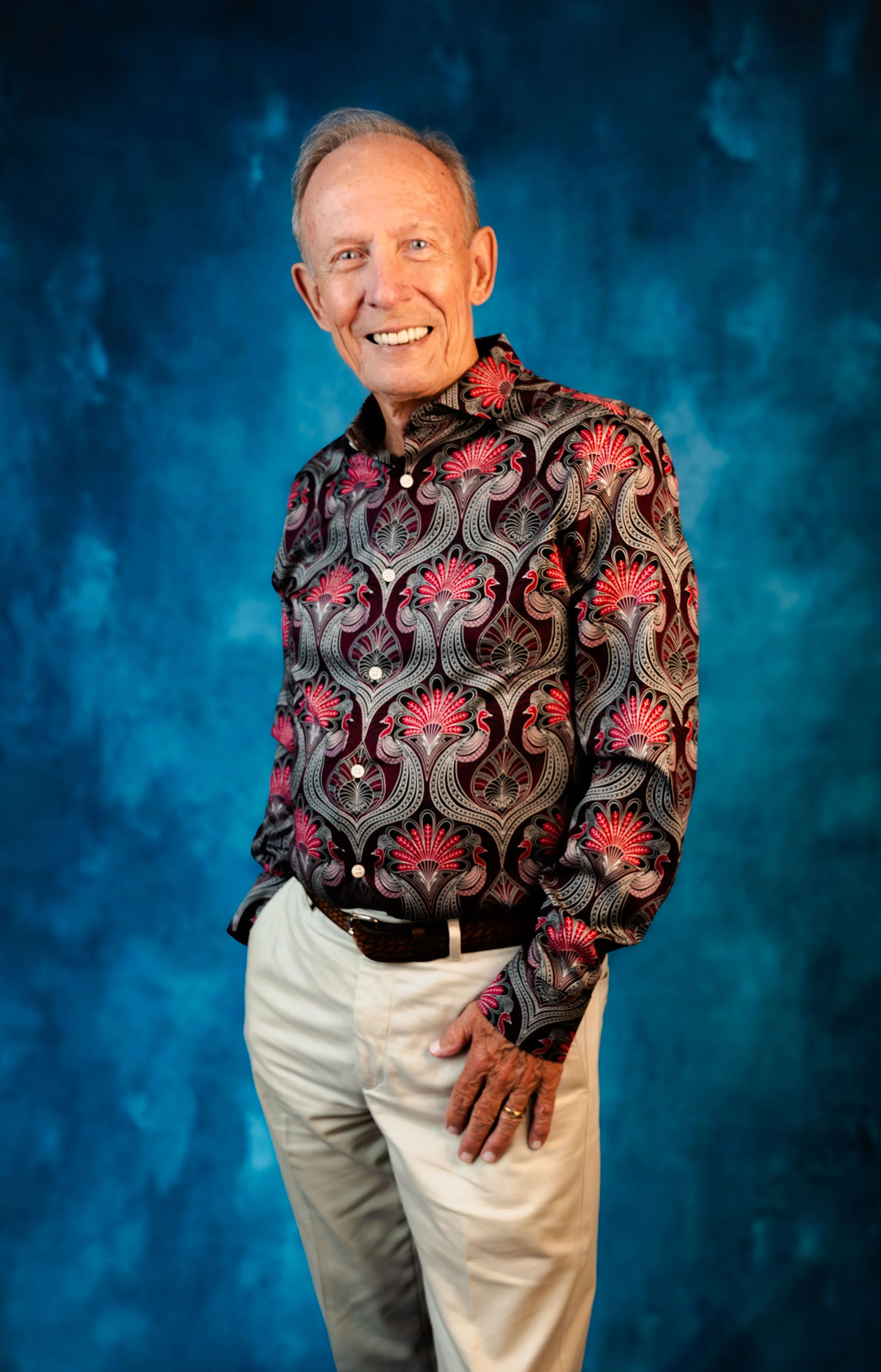 An elderly man with light skin, gray hair, and a bright smile, wearing a colorful patterned shirt with reds and blacks, beige pants, standing against a vibrant blue textured background.