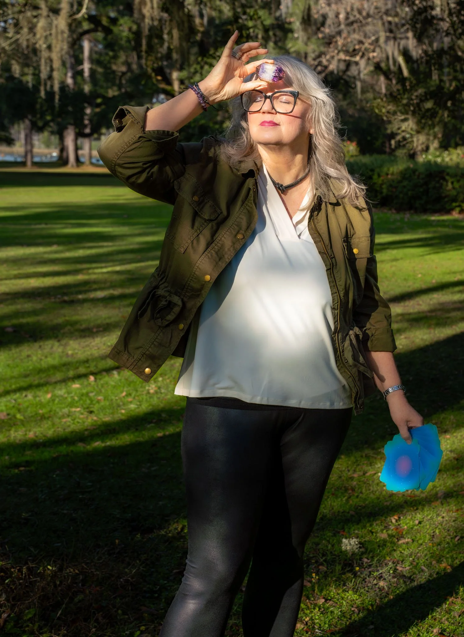 A woman with gray hair and glasses holding a purple crystal on her forehead outdoors in a park with green grass and trees.