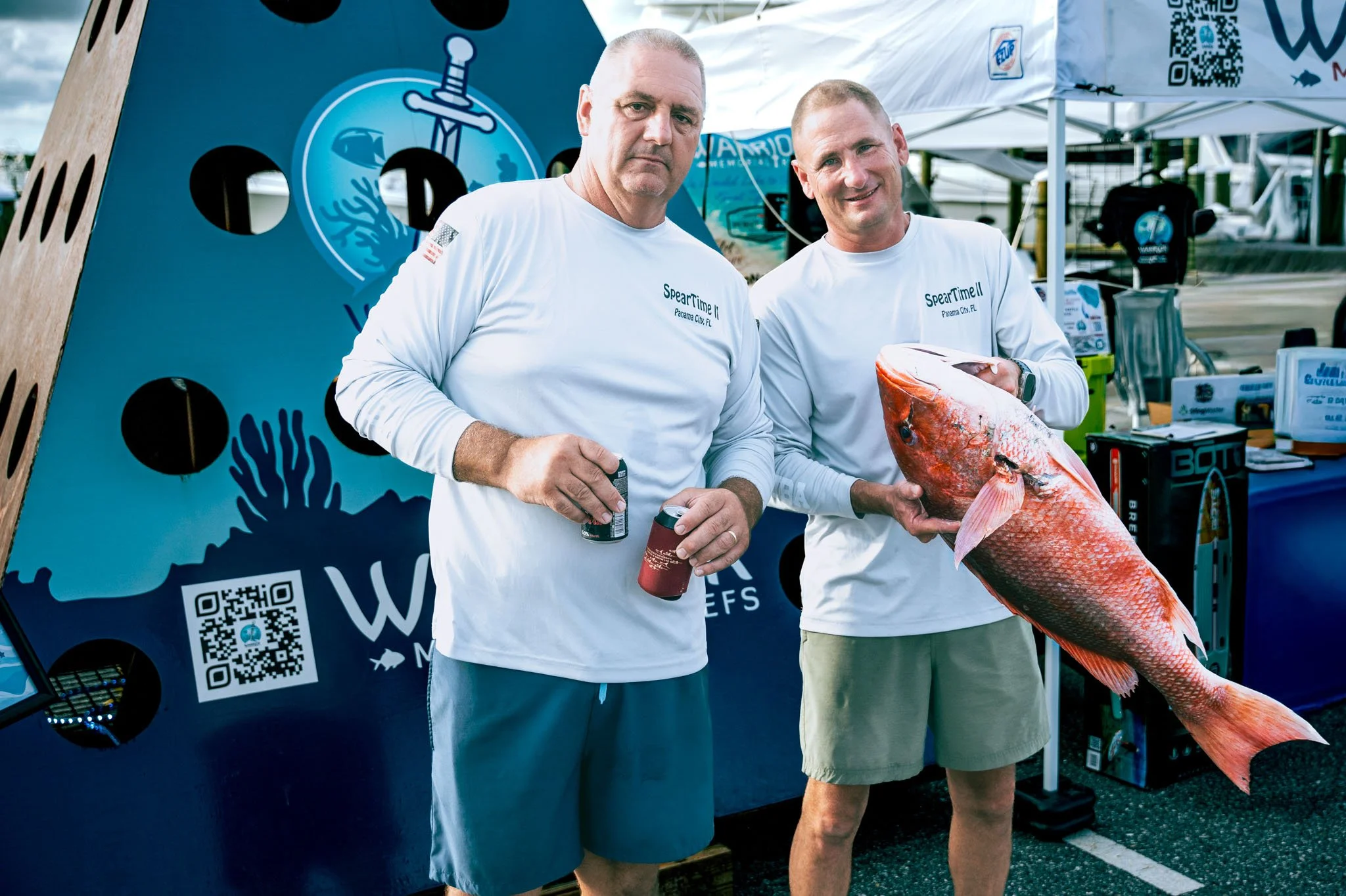 Two men wearing white shirts with 'Spear Time' and 'Panama City, FL' printed on them, standing outdoors at a booth or event with tents in the background. One man is holding a large red fish, and the other is holding canned drinks.