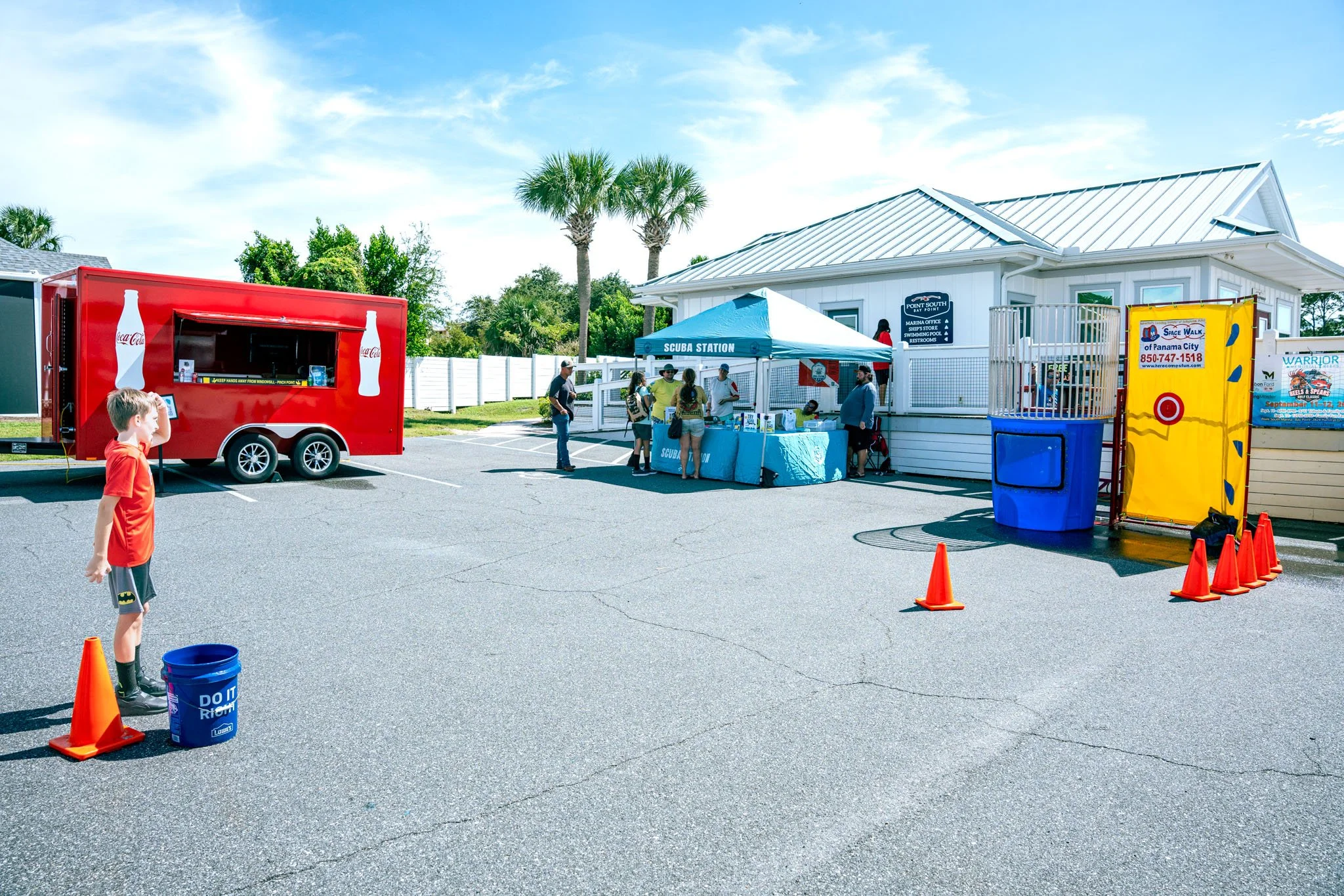 Point South Marina - Bay Point (Panama City Beach), people waiting in line at an outdoor booth under a blue canopy labeled 'Scuba Station,' with a white building in the background, a red Coca-Cola trailer, and children playing nearby on a sunny day.