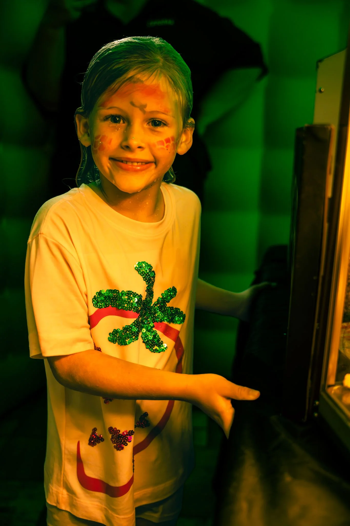 Young girl with face paint and a glittery green shamrock on her shirt, standing in front of a green background, smiling.