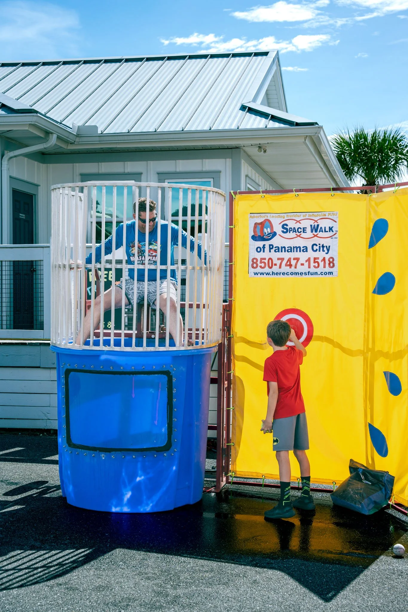 A man and boy playing a game at an outdoor carnival or fair. The man is sitting in a cylindrical blue container with a guardrail, and the boy is at a target on a yellow wall, holding a toy gun. The sign on the wall reads 'Space Walk of Panama City' w