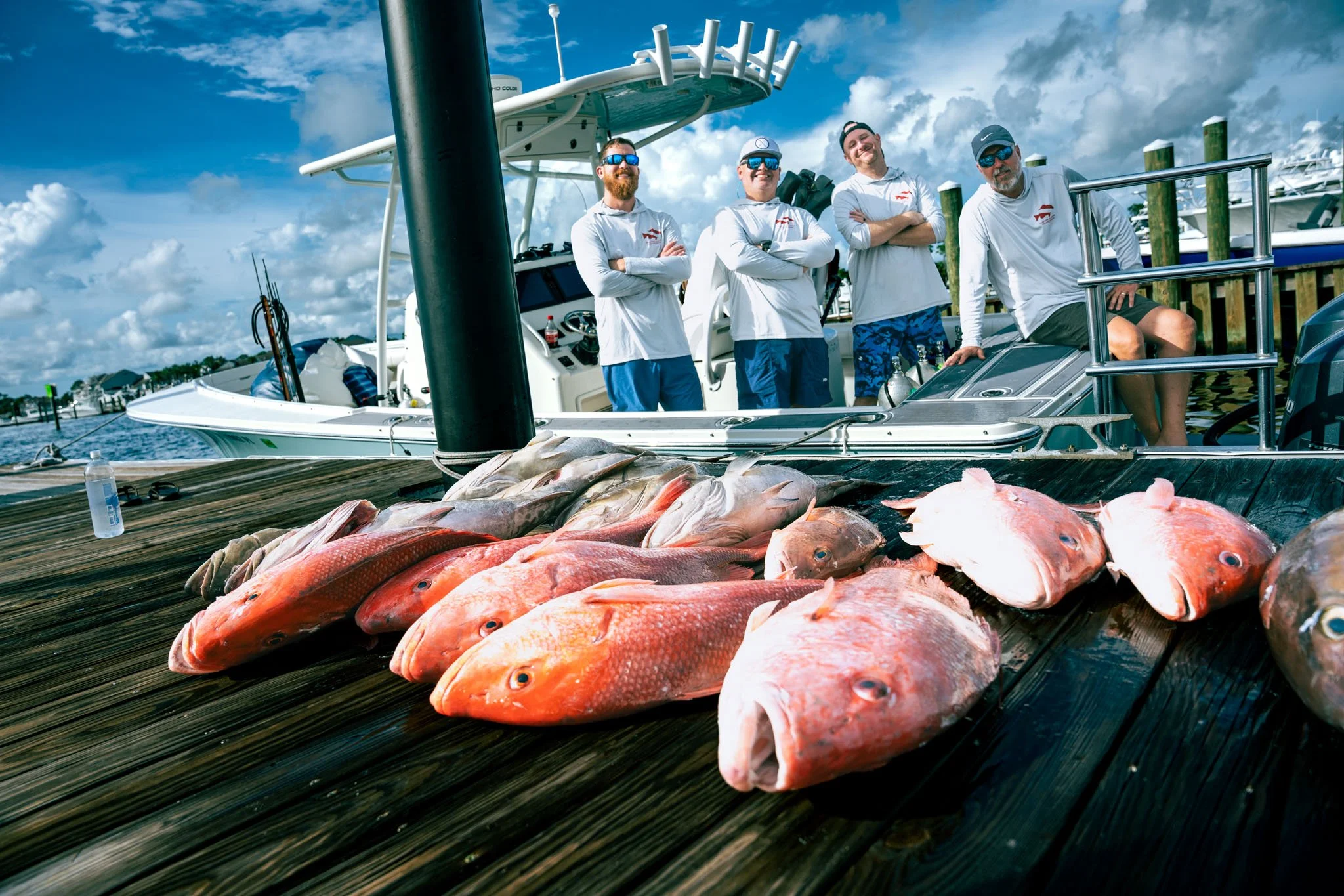 Four men in white shirts and sunglasses standing on a boat docked at Point South Marina - Bay Point (Panama City Beach), with a helicopter-style fishing boat behind them and fish laid out on a wooden dock in the foreground.