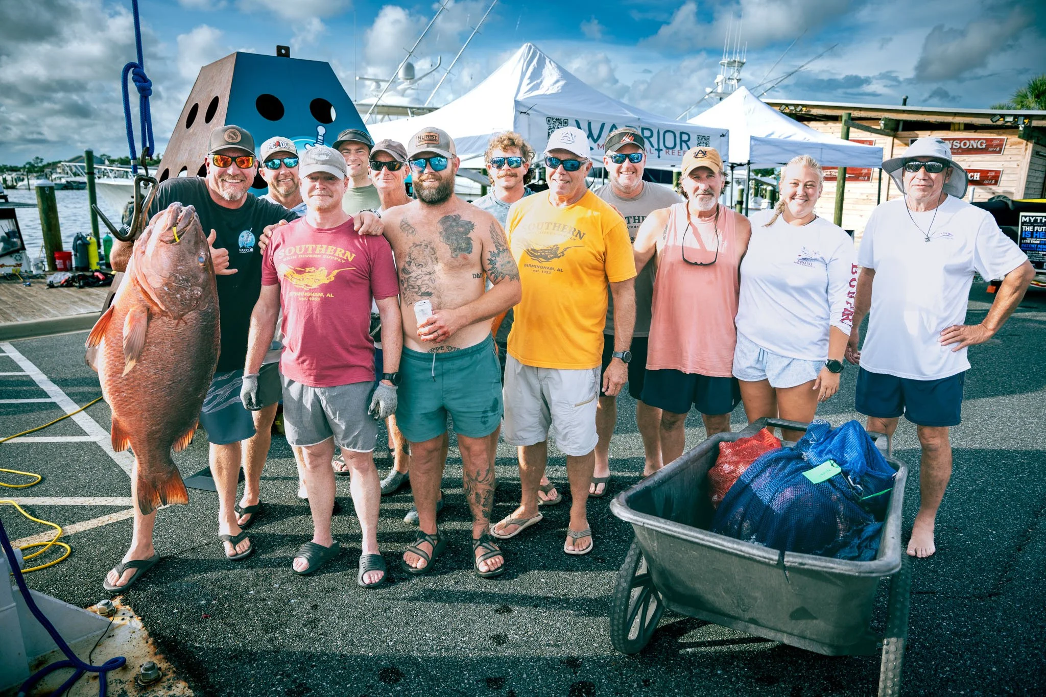 A group of people, mostly men, standing on a boat dock holding a large cubera snapper, with tents and boats in the background under a partly cloudy sky.
