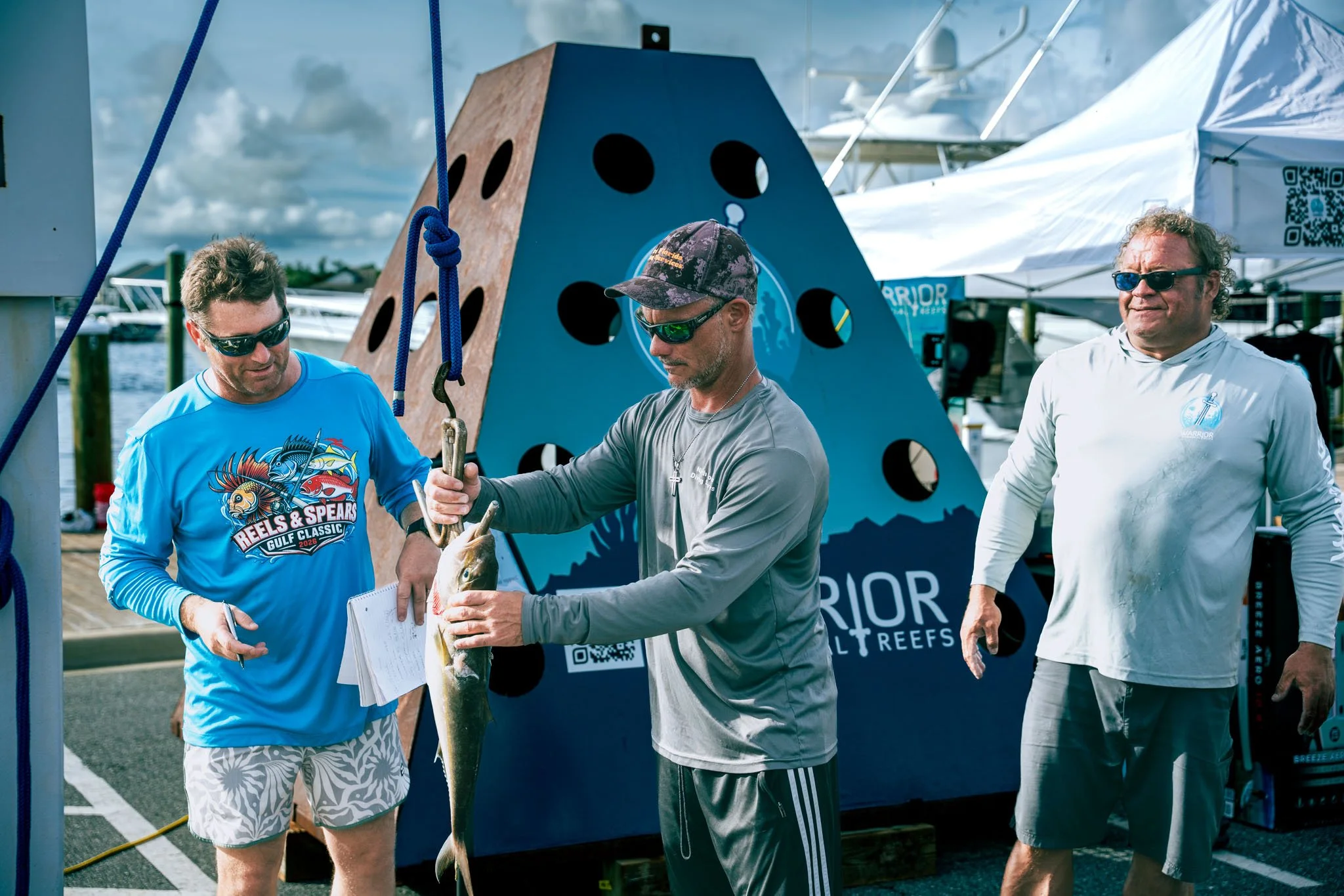 Three men at a marina during daytime, one holding a fish. The man on the left is wearing a blue long-sleeve shirt with colorful graphics and patterned shorts. The middle man, wearing a gray shirt and a camouflage cap, is examining the fish. The third