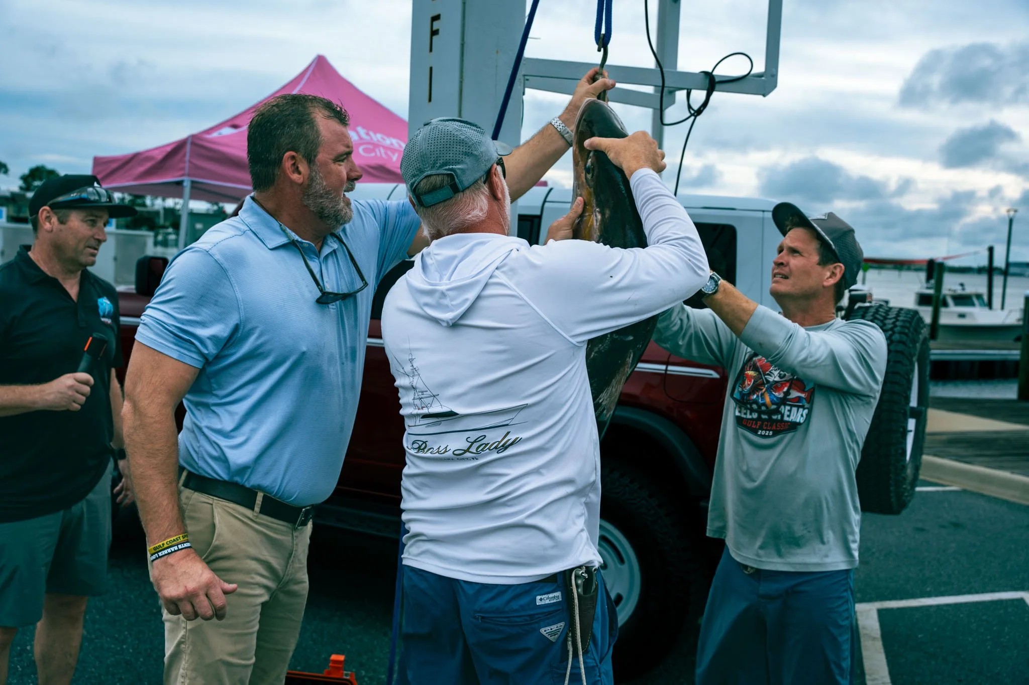 Four men are at a dock, with a pink tent and boats in the background. One man is holding a fish on a scale with assistance from another man. The group appears engaged in weighing the fish.