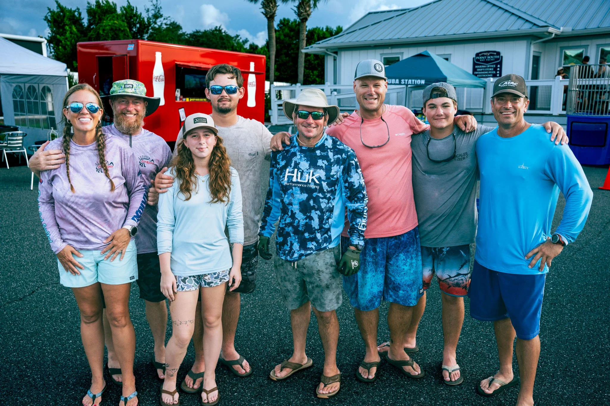 Group of nine people standing outdoors at a casual event, smiling, wearing sunglasses, summer casual clothing, with tents, a trailer, and a building in the background.