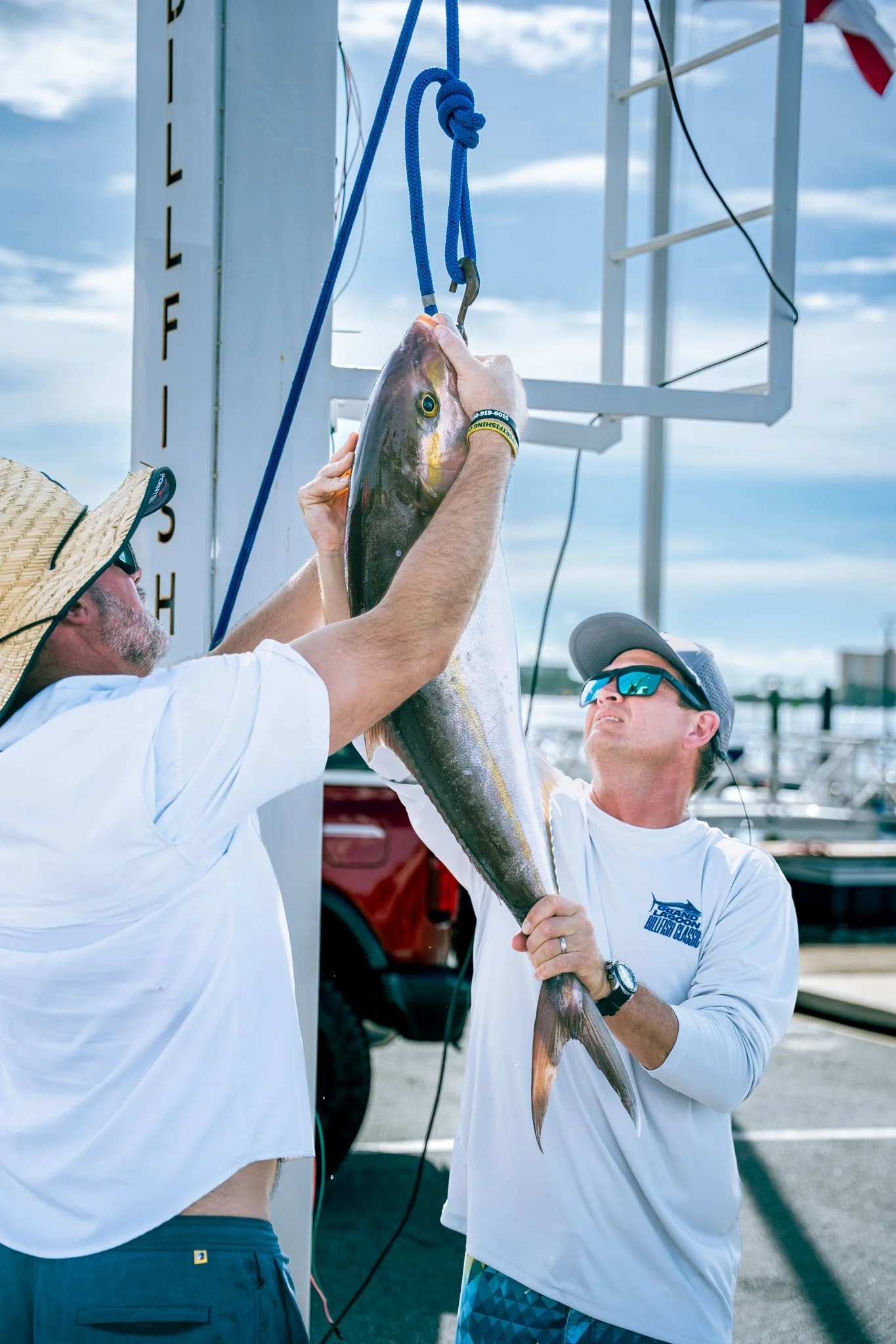 Two men in white shirts are holding a large fish, hanging from a blue strap, with a third person observing. They are outdoors near boats, with a partially cloudy sky in the background.