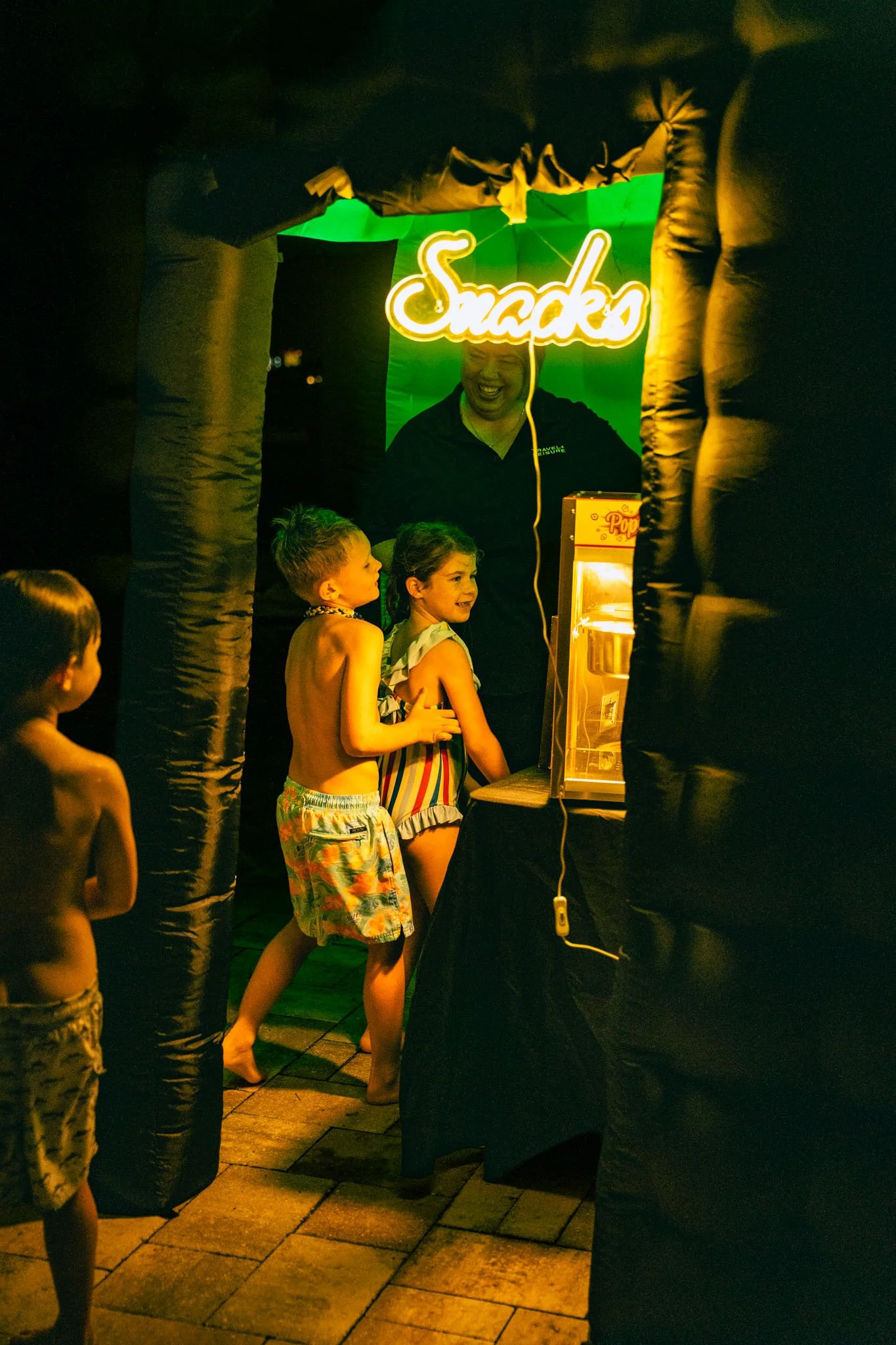 Children in swimwear standing in line at a nighttime fair, waiting for popcorn, with a lit sign that says "Snacks" in the background.
