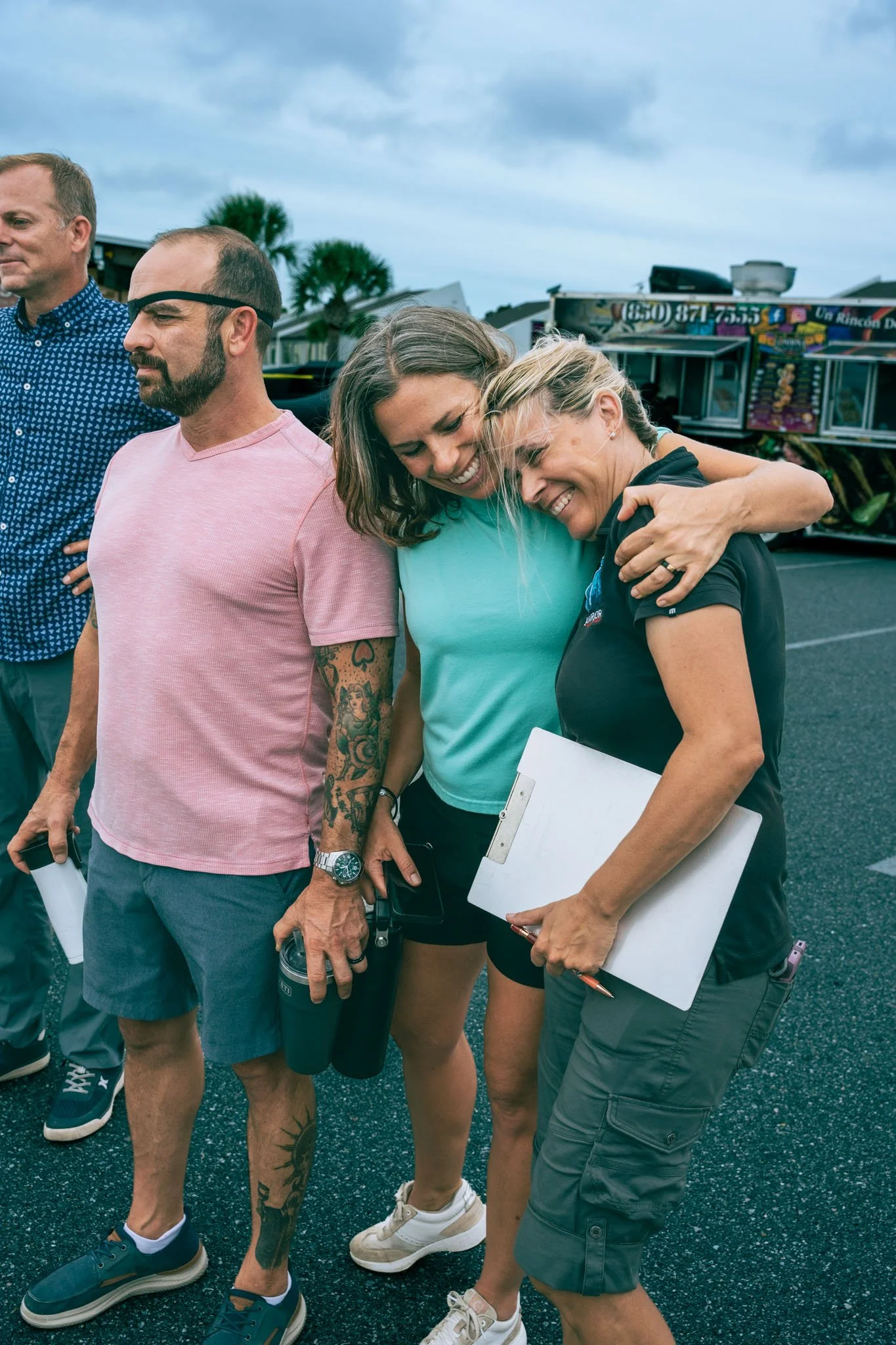 A group of five people standing closely together outdoors at Point South Marina - Bay Point (Panama City Beach), smiling and hugging. There is a food truck in the background on a cloudy day.