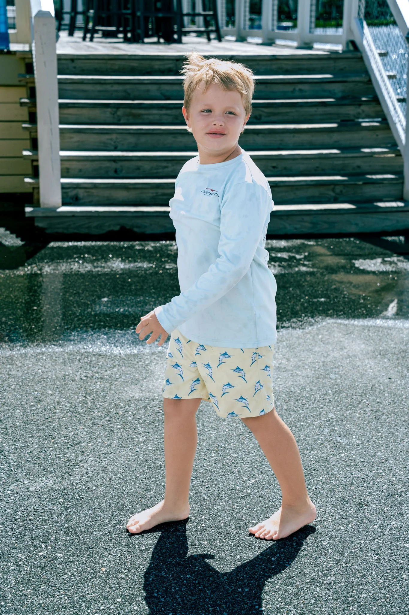 Point South Marina - Bay Point (Panama City Beach), a young boy with messy blond hair standing barefoot on a wet asphalt surface, wearing a long-sleeve white shirt and beige shorts with a blue shark pattern, with a wooden staircase in the background.