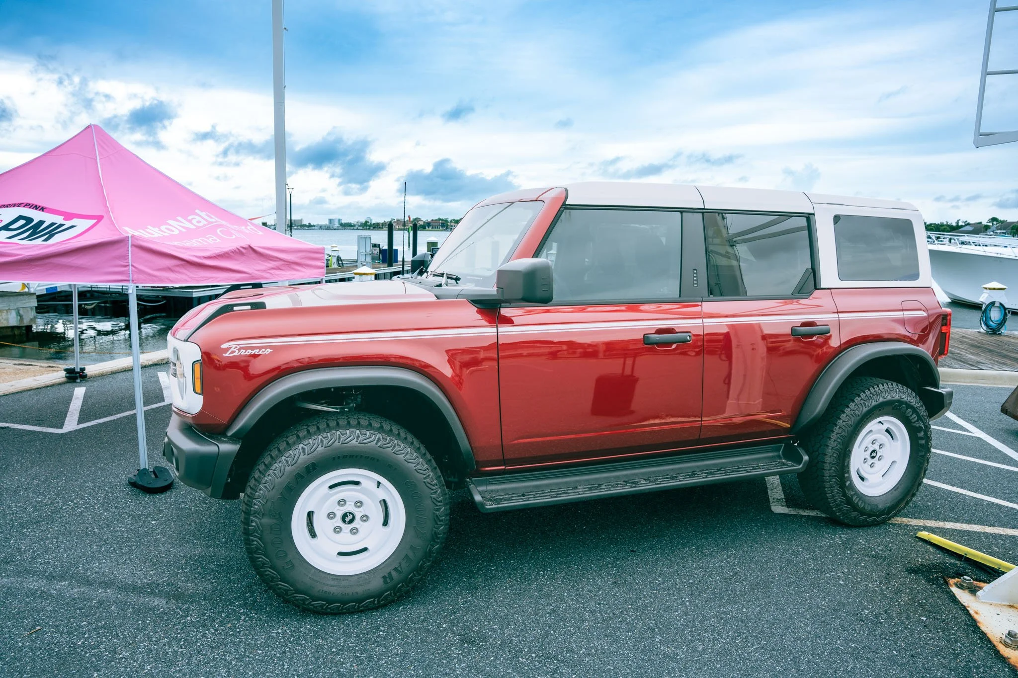 Red off-road Bronco parked at Point South Marina - Bay Point (Panama City Beach) next to a pink tent with an auto dealer logo, water and boats in the background, under a cloudy sky.