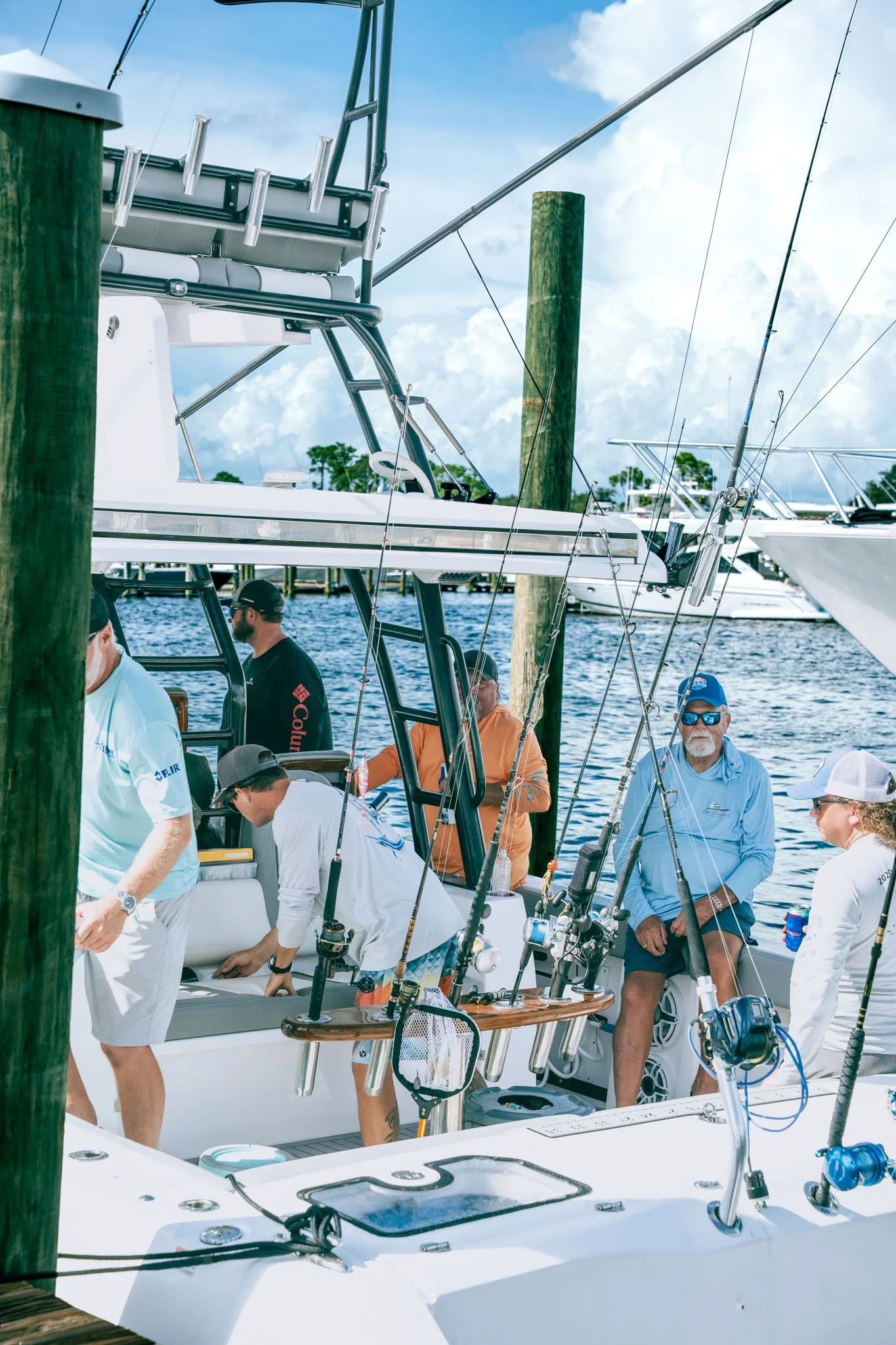 People on a boat docked in at Point South Marina - Bay Point (Panama City Beach), some sitting and some standing, with fishing rods, equipment, and a wooden table on the boat. The background shows water, other boats, and a partly cloudy sky.