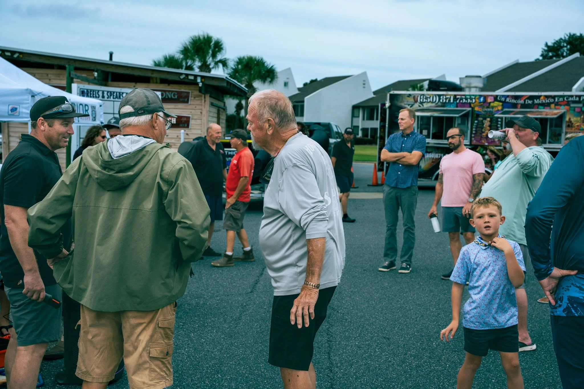 A group of people gathered outdoors at a fair or festival, with food trucks and buildings in the background. Some individuals are talking, and a young boy is looking at the camera.