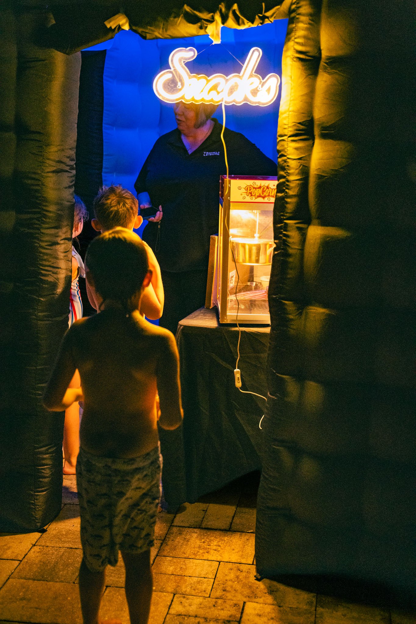 Children watching a performer at a carnival or fair, with a popcorn stand in the background and a neon sign that says 'Snacks'.