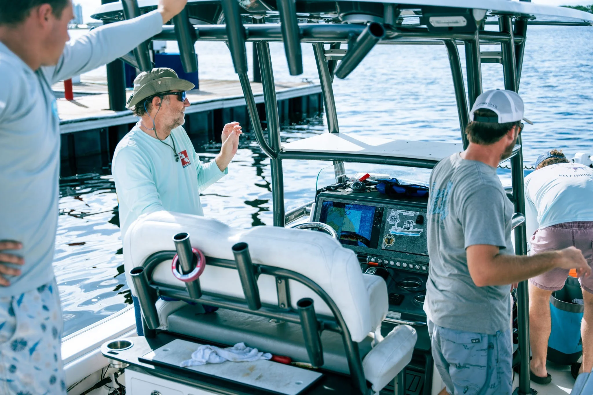 Group of people on a boat near the water, with one person wearing a hat and sunglasses, standing next to the boat's electronic equipment, while others are gathered nearby.
