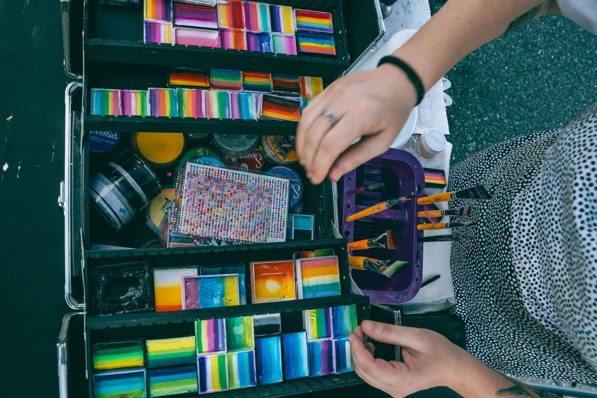 Open case containing various face paints, brushes, and art supplies on a white table, with a person arranging the paints.