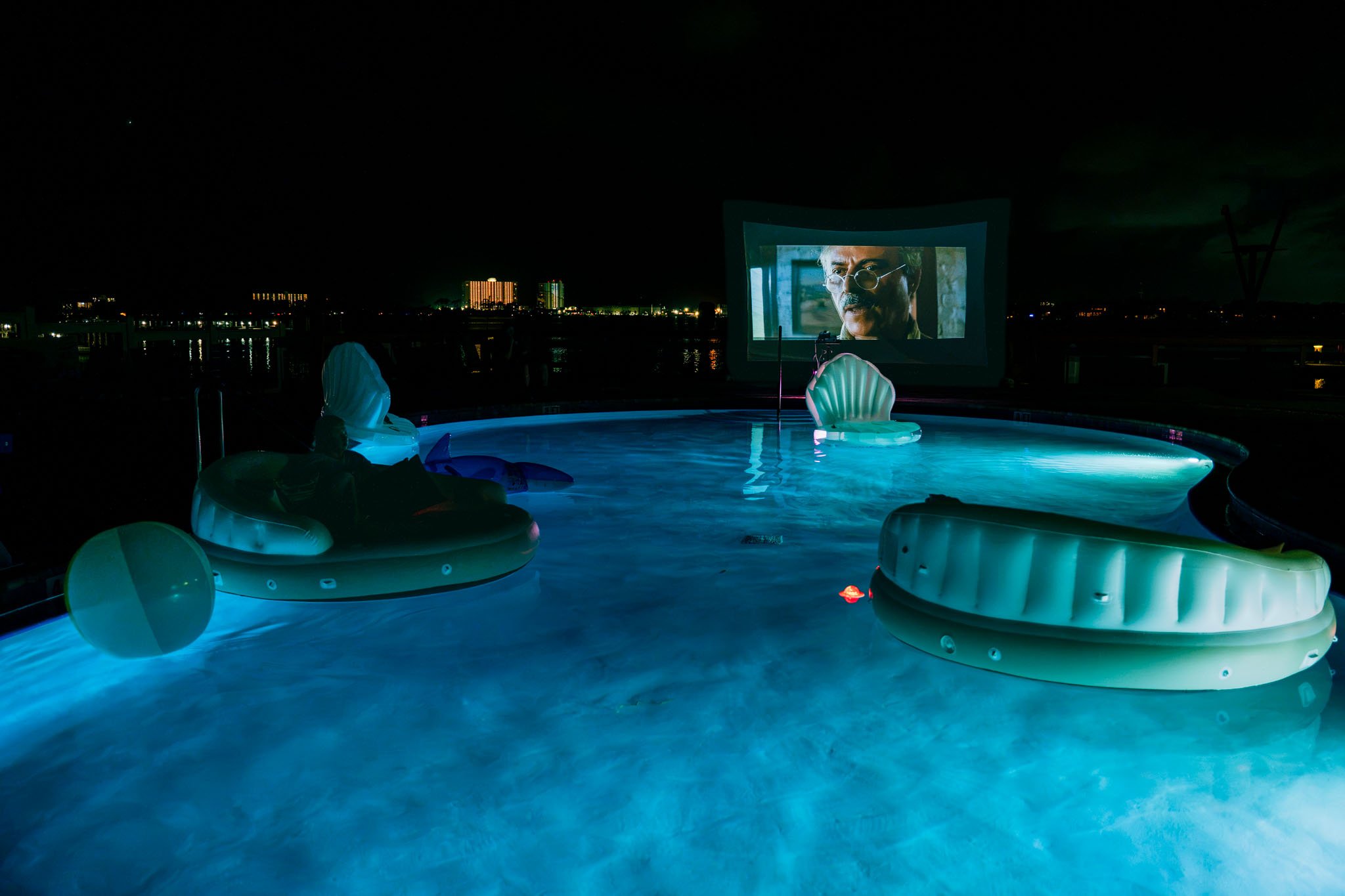 Nighttime view of Point South Marina - Bay Point (Panama City Beach) swimming pool with glowing inflatable pool floats, a large screen showing a man's face, and city lights in the background.