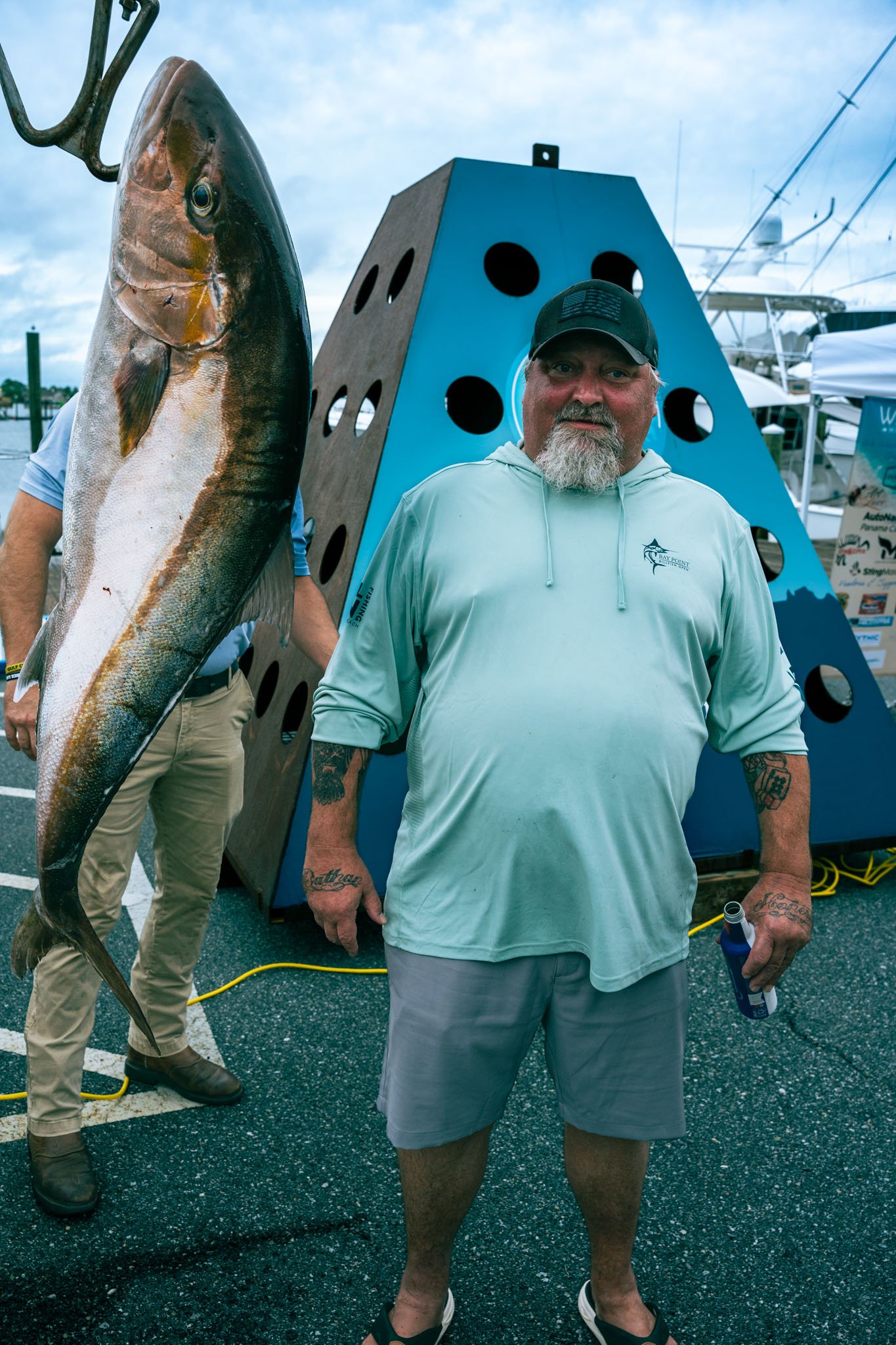 A man with a beard, wearing a cap and a hoodie, standing on a dock holding a fishing rod in one hand and a beer in the other. There is a large fish hanging on his left side and a blue structure with holes behind him. The background shows boats and a 