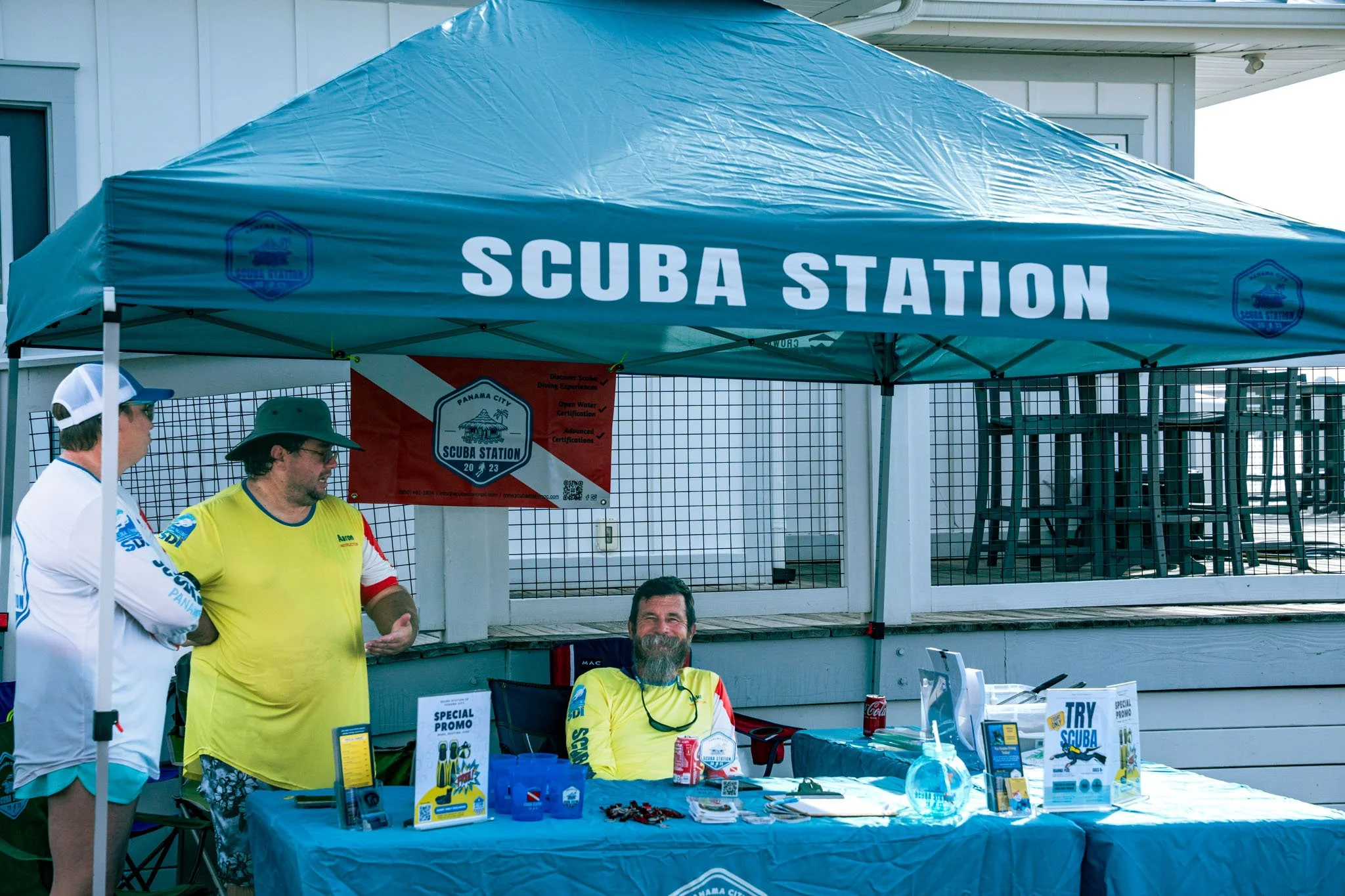 People standing and sitting at a booth with a blue canopy labeled "Scuba Station" during daytime, with promotional materials and drinks on the table.
