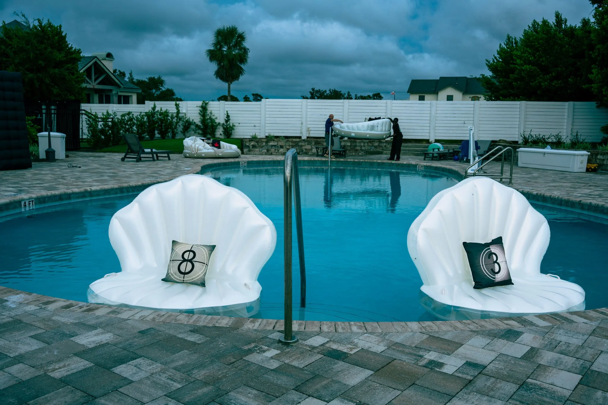 Two white pool chairs with countdown pillows, a swimming pool, pool toys, and two people setting up a float in an outdoor backyard pool area on a cloudy day.