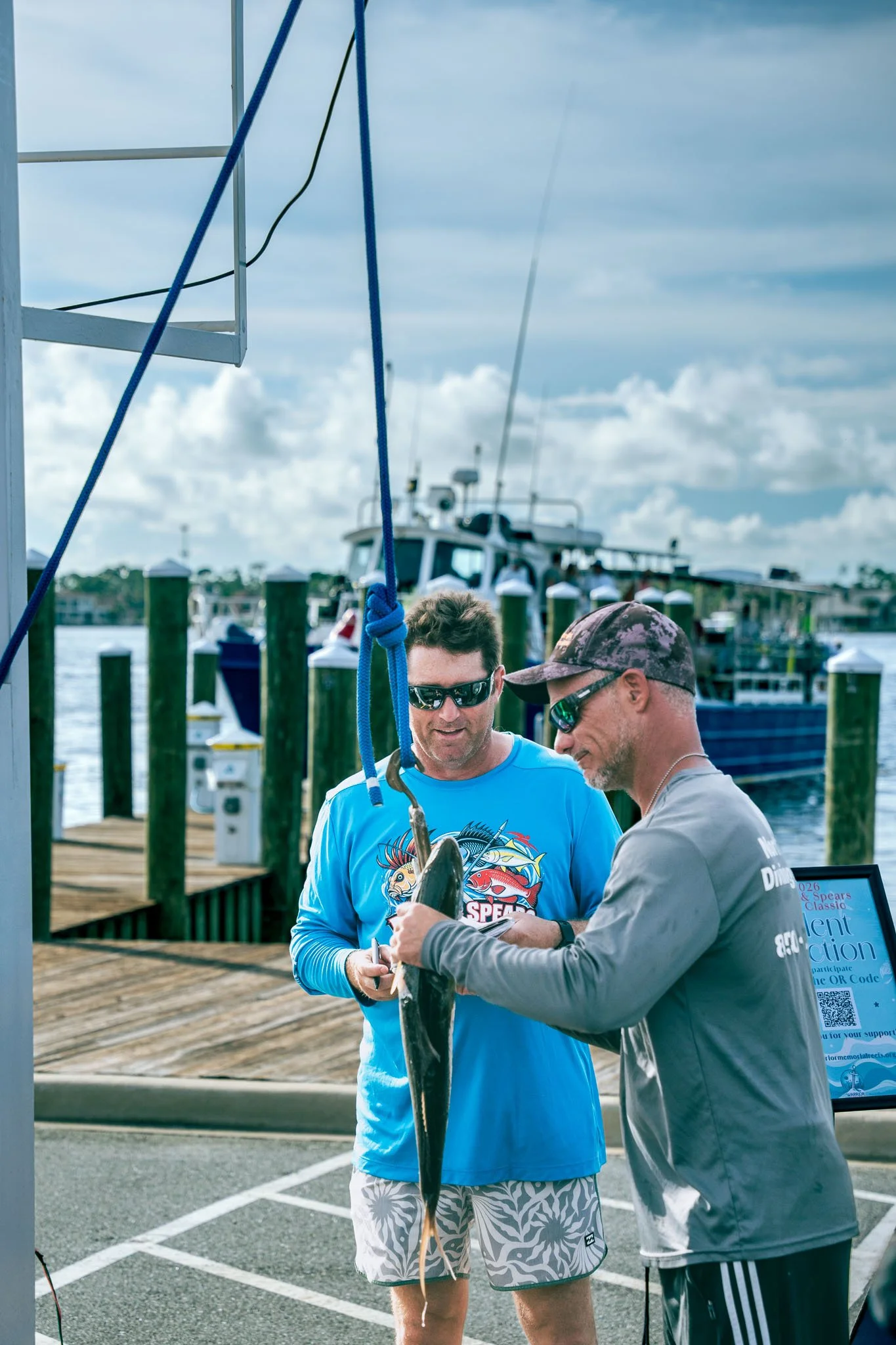 Two men on a dock at Point South Marina - Bay Point (Panama City Beach) show a gag grouper to each other, with boats and a cloudy sky in the background.