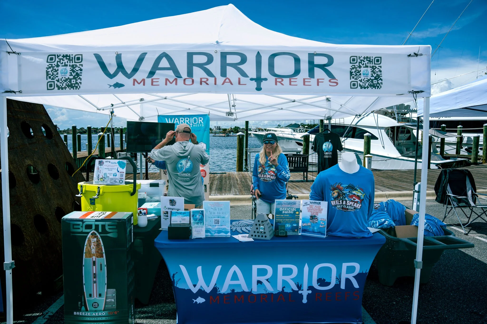 A canopy with the words "WARRIOR MEMORIAL REEFS" and a QR code on each side. Underneath, two people are smiling; one man is adjusting his hat, and one woman is holding her hand up in a peace sign. The booth has promotional materials, T-shirts, and a 