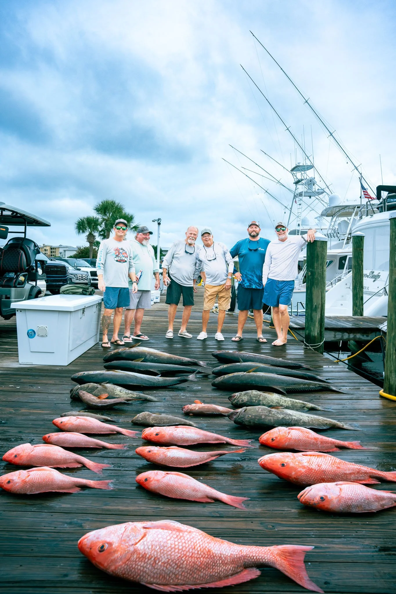 Group of five men standing on a dock with freshly caught fish laid out in front of them, boats docked in the background, cloudy sky overhead.