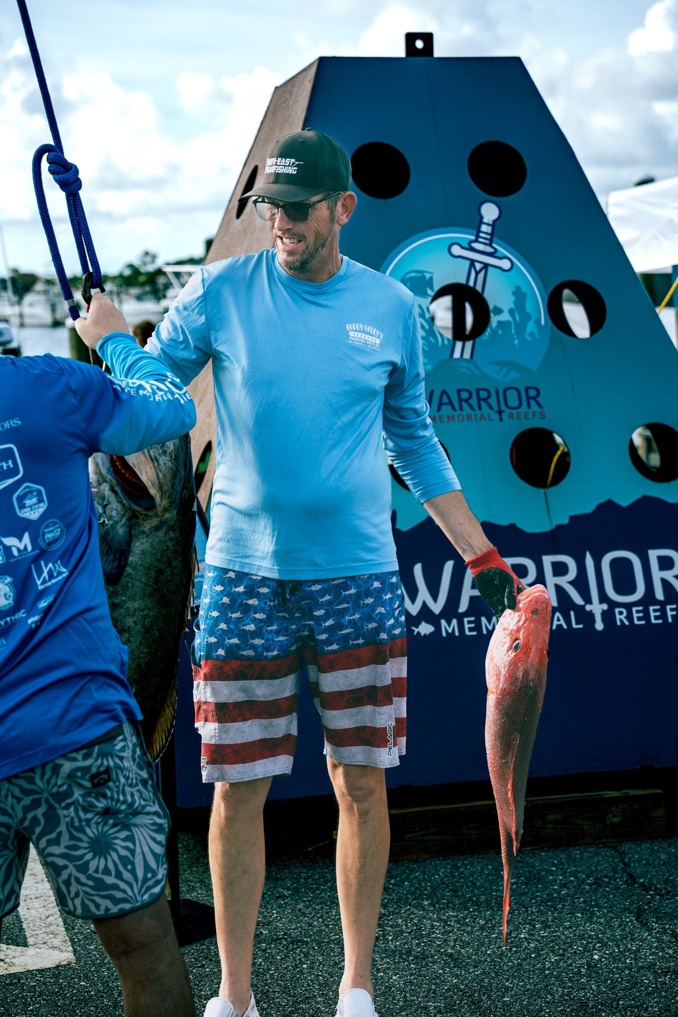 Man in striped swim trunks holding a red fish in front of a blue Warrior Memorial sign, with another person partially visible in front of him holding a large fish.