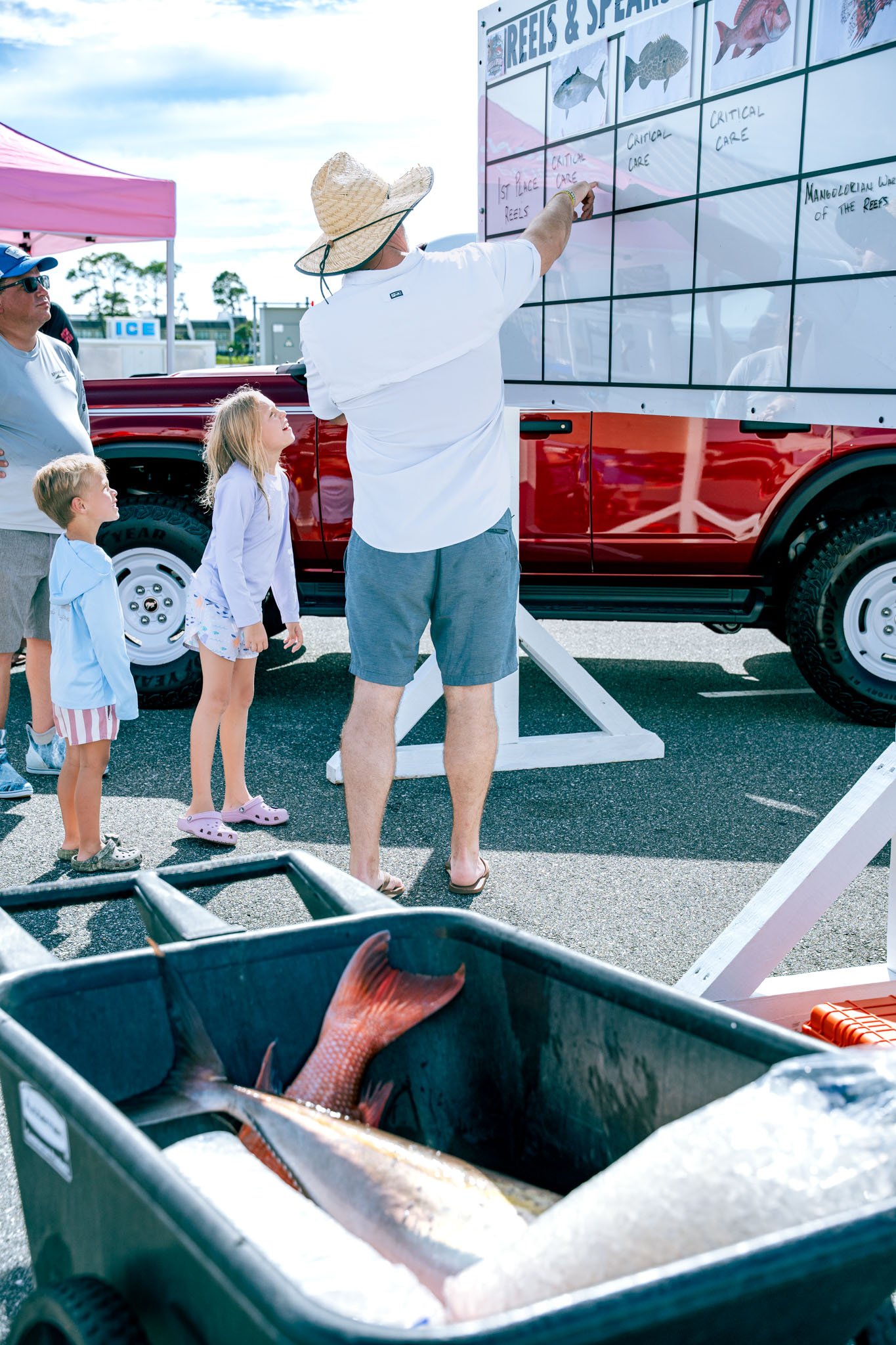 A group of people, including a man in a large sun hat and shorts, two children, and a woman, are gathered around a whiteboard outdoors. The whiteboard has drawings of fish and categorizes them as reef fishes under various care categories. In the fore